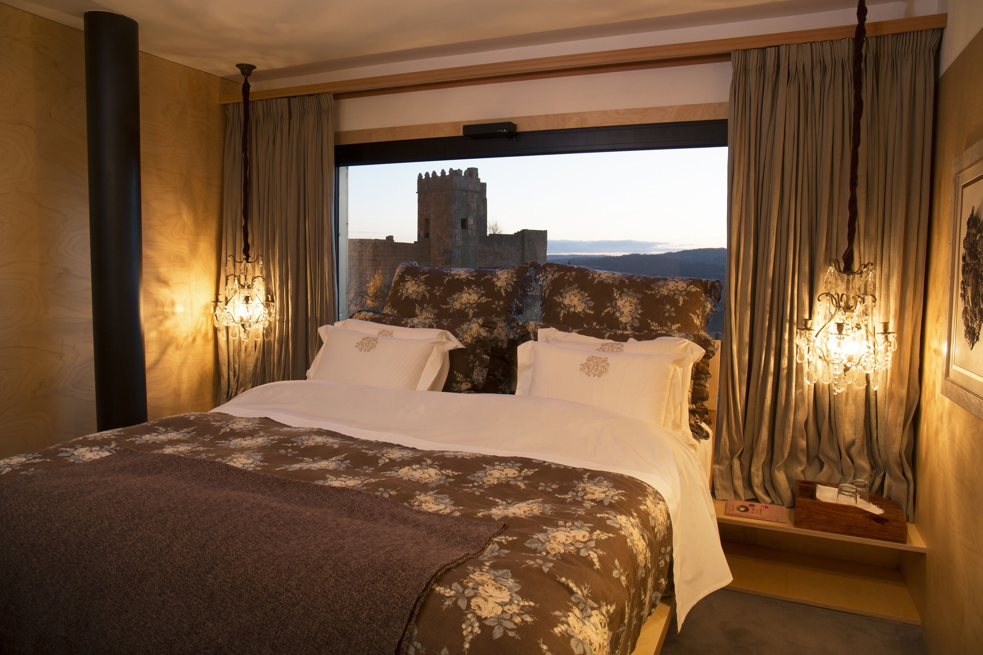 Un dormitorio con una cama grande, ropa de cama floral, vistas de un castillo a través de una ventana y dos lámparas fijadas a la pared.
