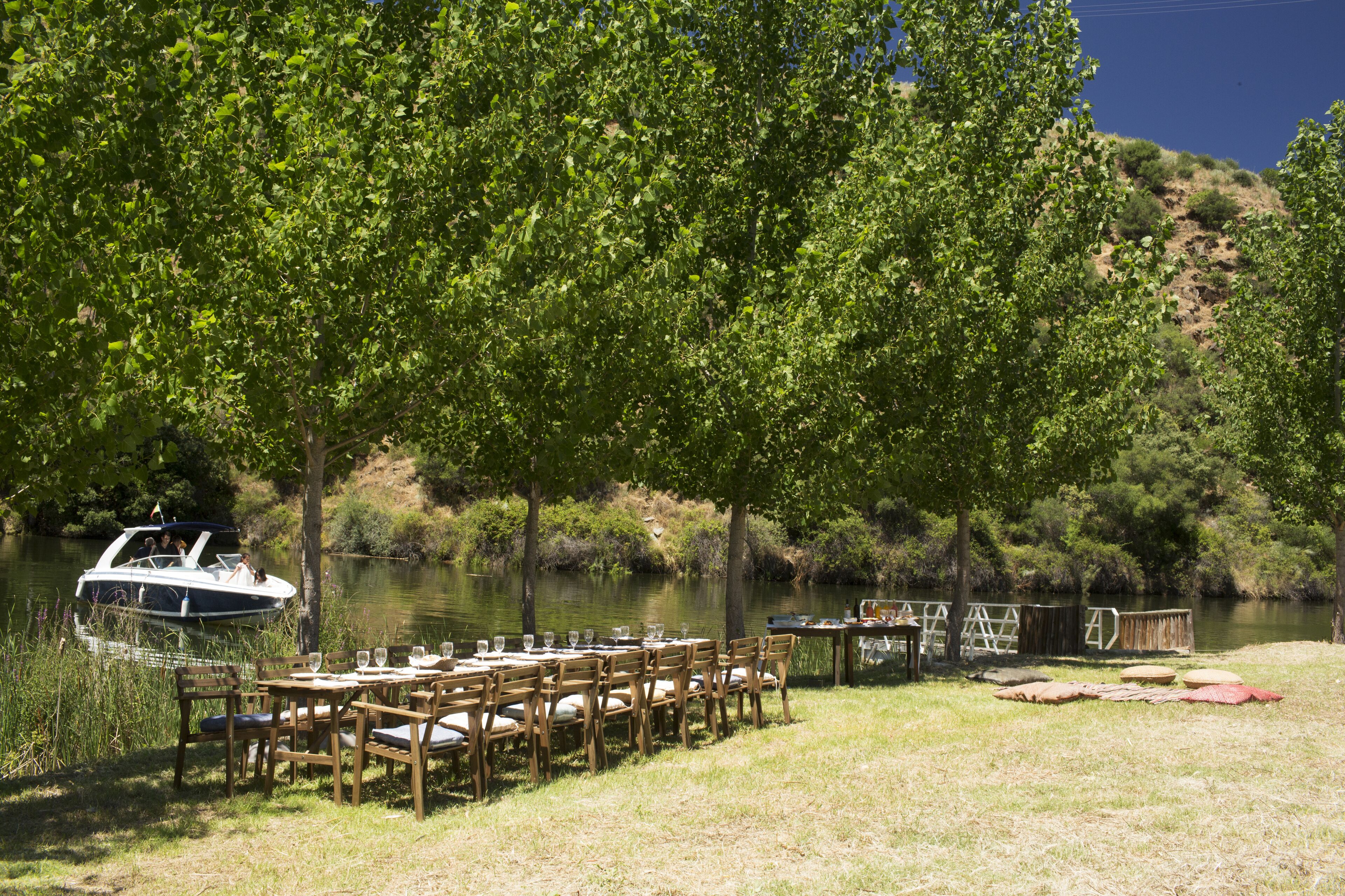 An outdoor dining area with tables and chairs set up near a body of water, a boat docked nearby, and a small structure in the background.