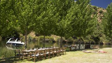 An outdoor dining area with tables and chairs set up near a body of water, a boat docked nearby, and a small structure in the background.