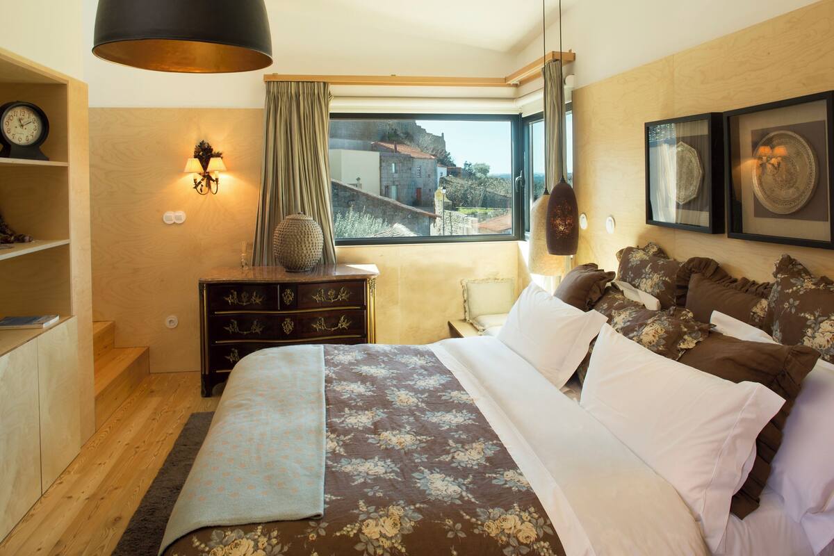 A bedroom with a large bed, a dresser, a window with a view, and a wall-mounted lamp.