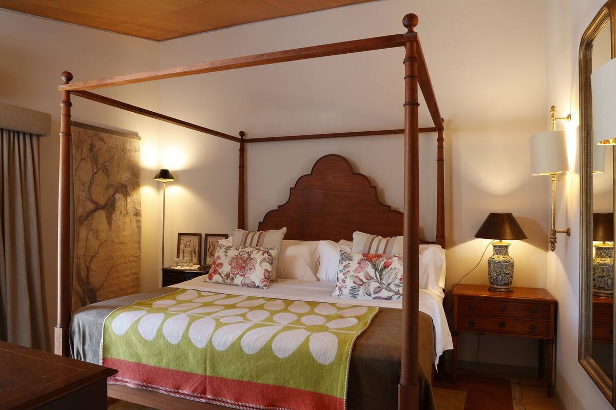 A four-poster bed with a floral bedspread, a nightstand with a lamp, and a mirror on the wall.