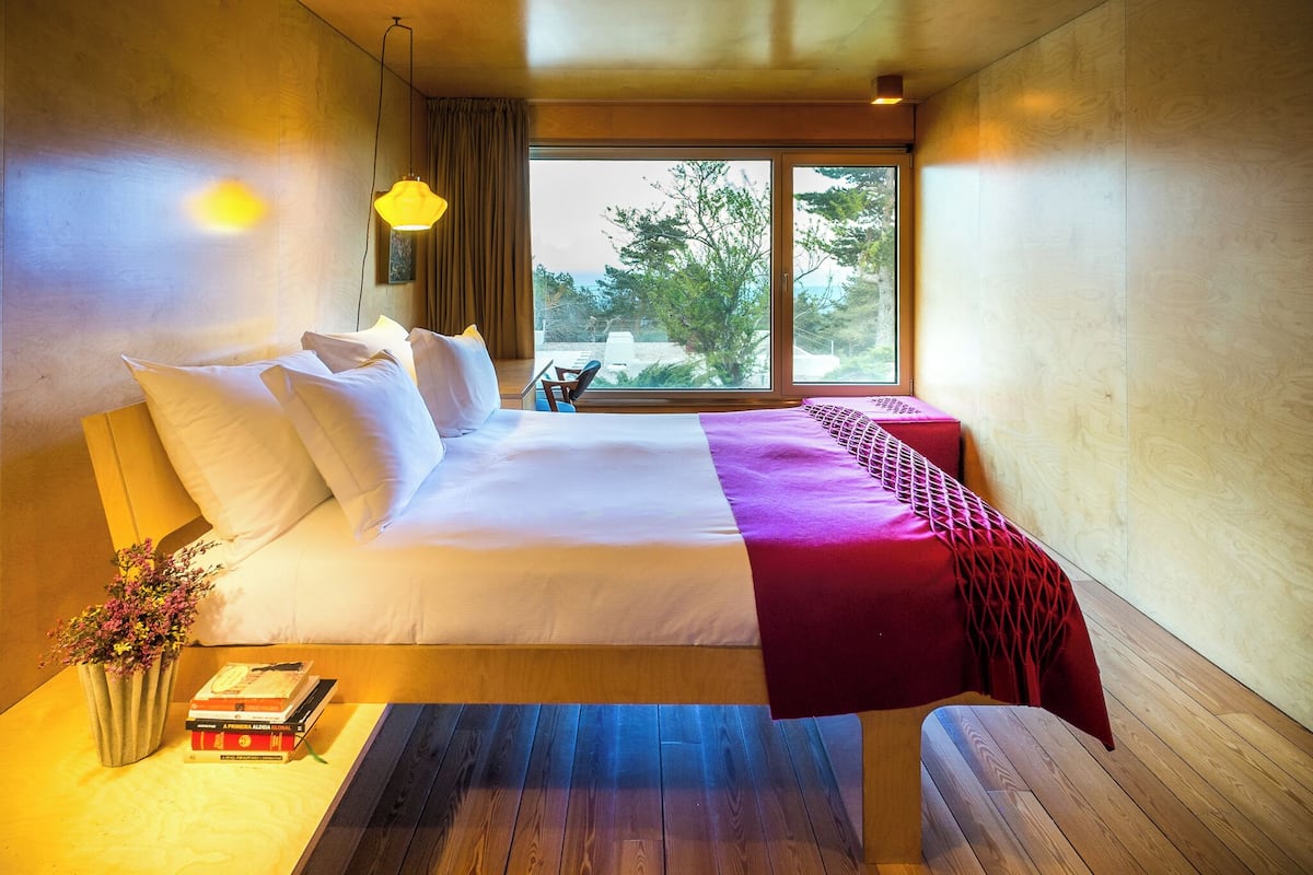 Ein modernes Hotelzimmer mit einem großen Bett, einem Nachttisch mit Büchern, einem kleinen Tisch mit einem Sessel und einem großen Fenster mit Blick auf Bäume.