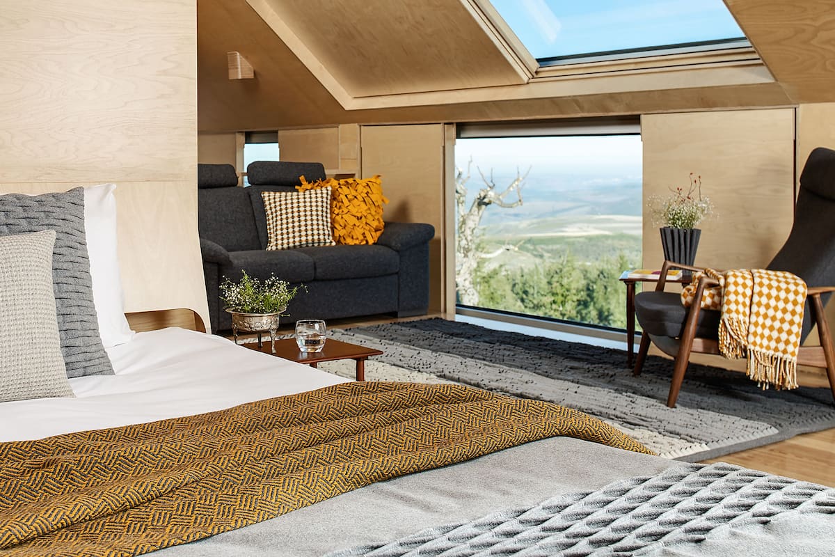 Ein modernes Zimmer mit Bett, Sofa und Sessel, einem großen Fenster mit Ausblick und einem Dachfenster.