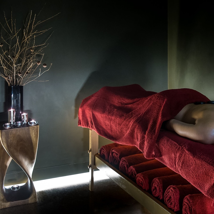 A massage therapy room with a wooden massage table, red towels, and a decorative vase with branches.