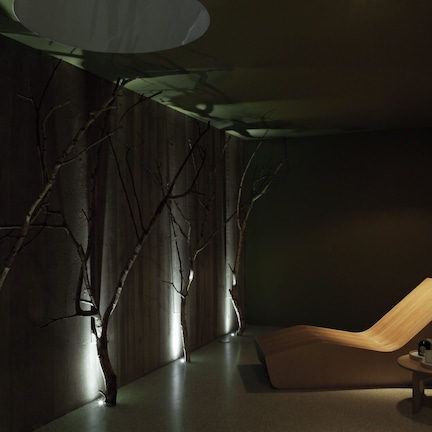A modern room with a wooden chair, a small table with a teapot, and a wall with branches.