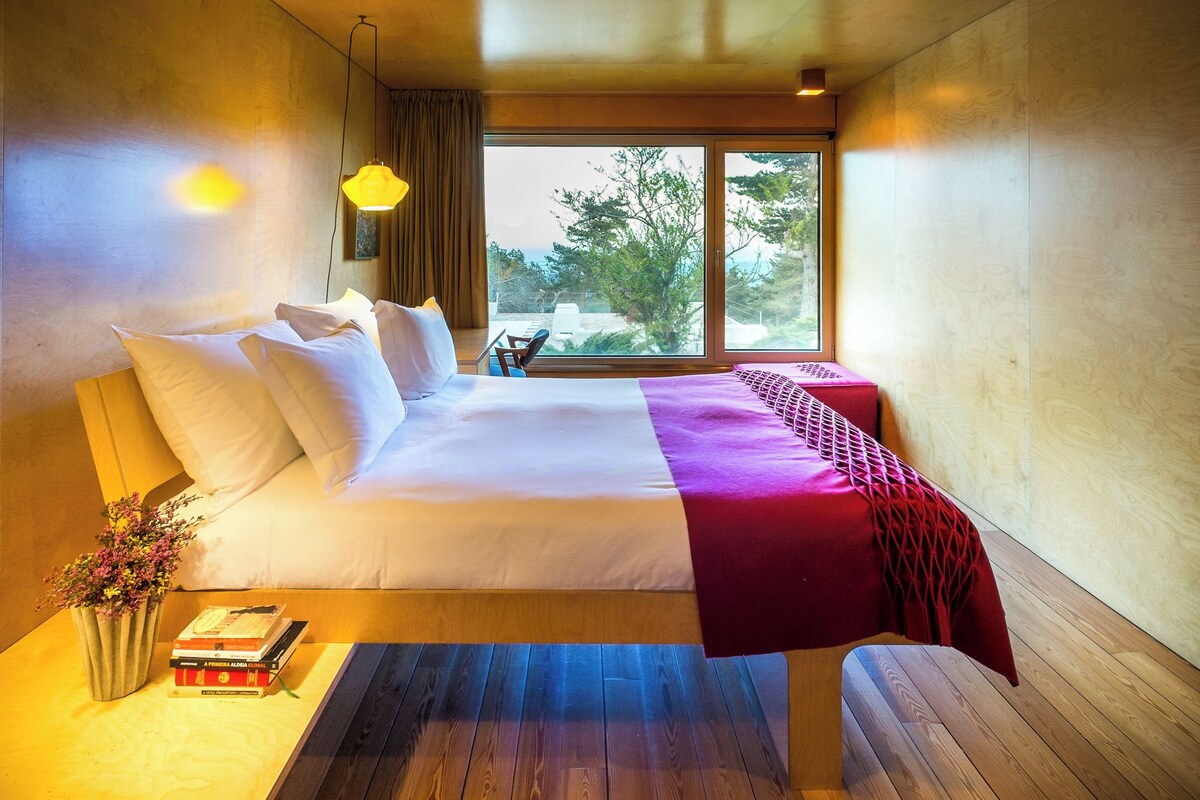 Ein modernes Hotelzimmer mit einem großen Bett, einem Nachttisch mit Büchern, einem kleinen Tisch mit einem Sessel und einem großen Fenster mit Blick auf Bäume.