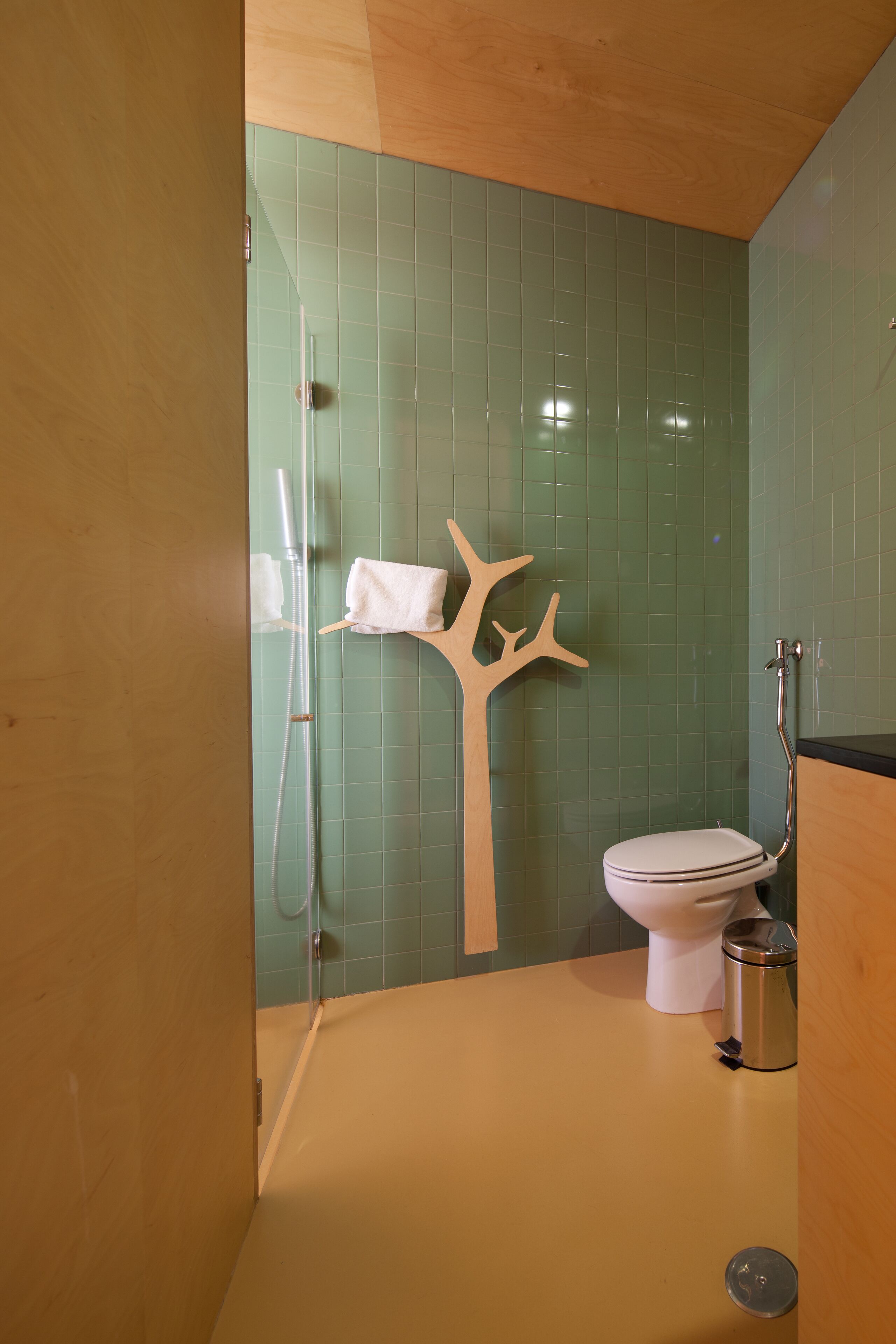 Een badkamer met een groene betegelde muur, een wit toilet, een douche met een glazen deur en een houten handdoekhouder in de vorm van een boom.