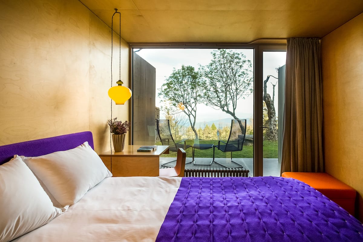 Ein Schlafzimmer mit einem großen Bett, Blick ins Freie und einem kleinen Tisch mit Lampe.