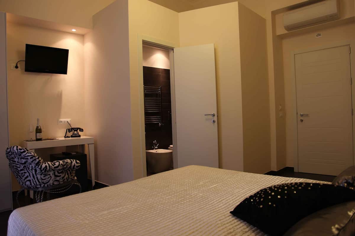 A hotel room with a bed, a desk with a chair, a television mounted on the wall, a bathroom with a sink and towel rack, and a door.