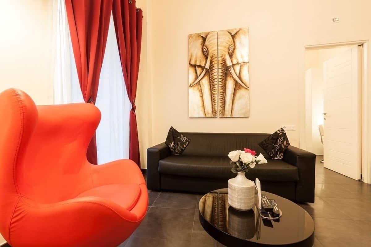 A modern living room with a black sofa, an orange chair, a coffee table with a vase of flowers, and a large elephant artwork on the wall.