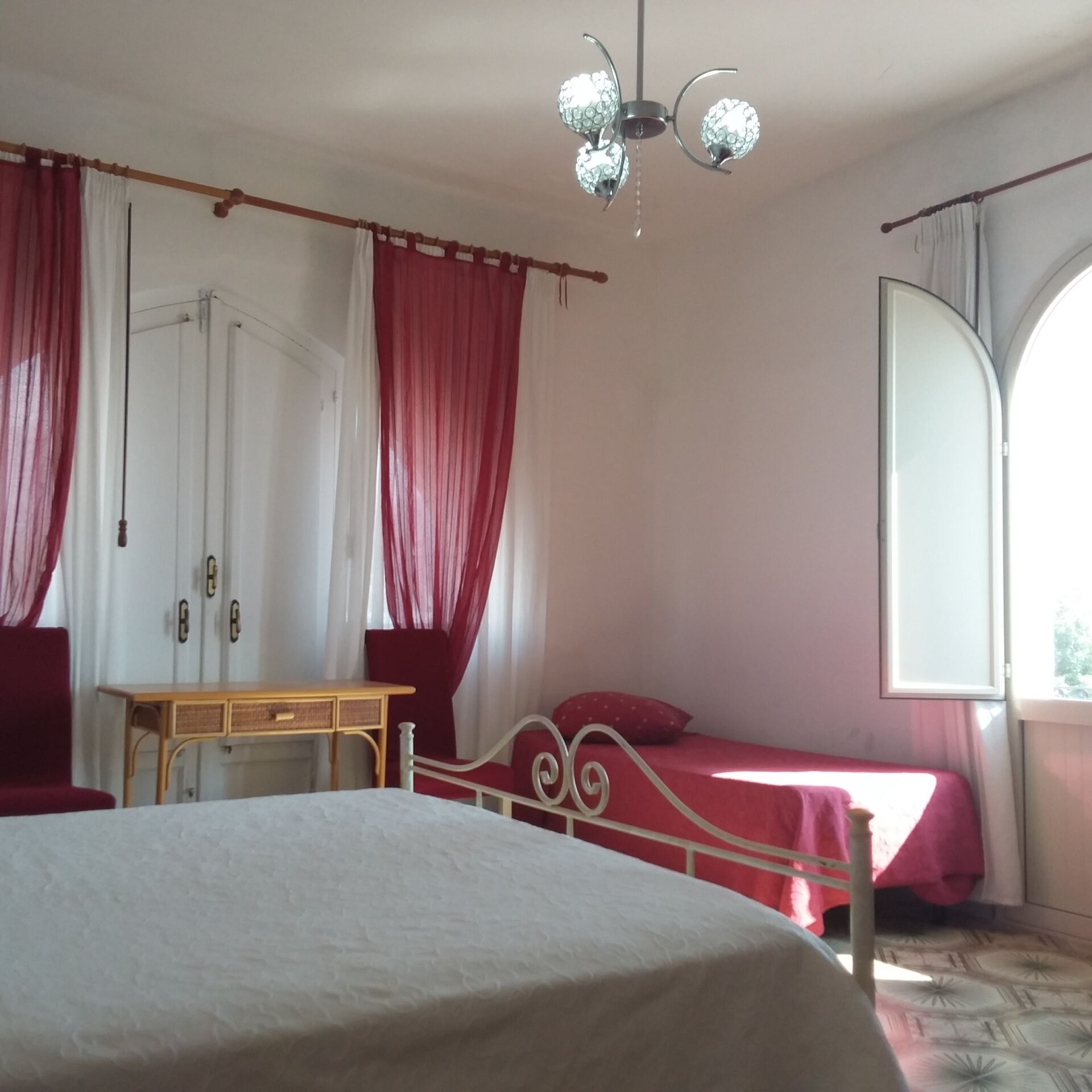 Una camera da letto con un letto, una scrivania e una sedia. Si vede una finestra con tende e un lampadario.