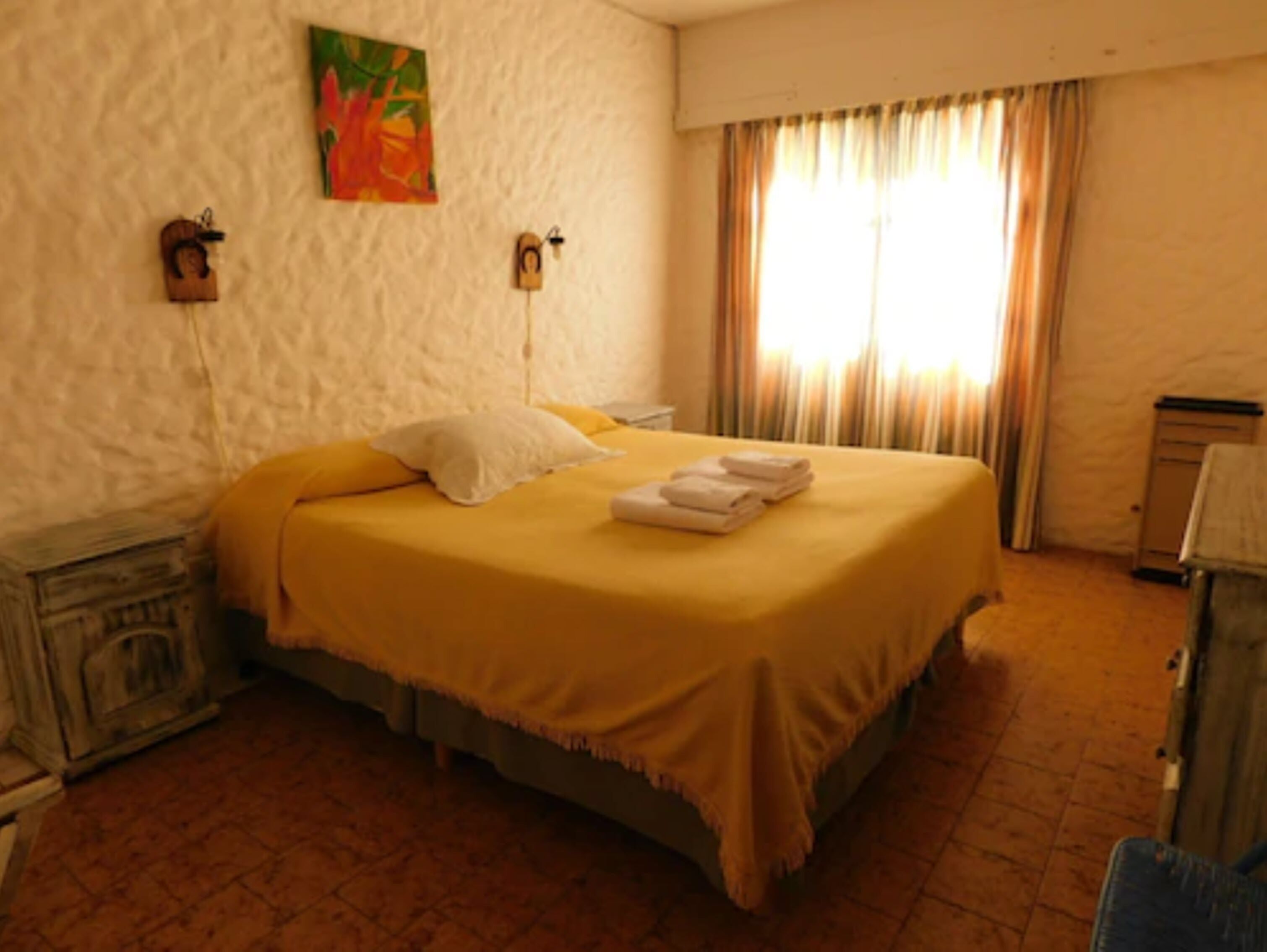 Una habitación de hotel con una cama, una mesita de noche, una lámpara, un cuadro y una ventana con cortinas.