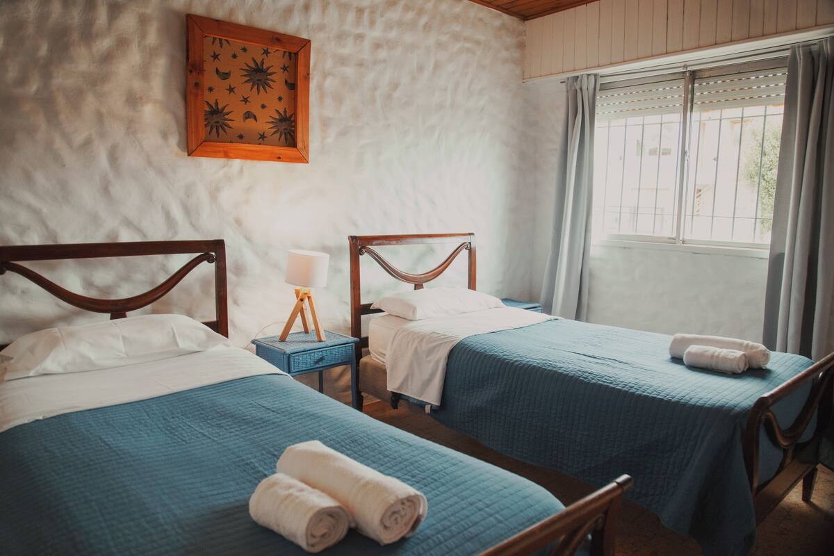 Une chambre avec deux lits, chacun doté d’une tête de lit en bois et d’une literie bleue. On y trouve une petite table avec une lampe, un tableau encadré au mur et une fenêtre avec des rideaux.