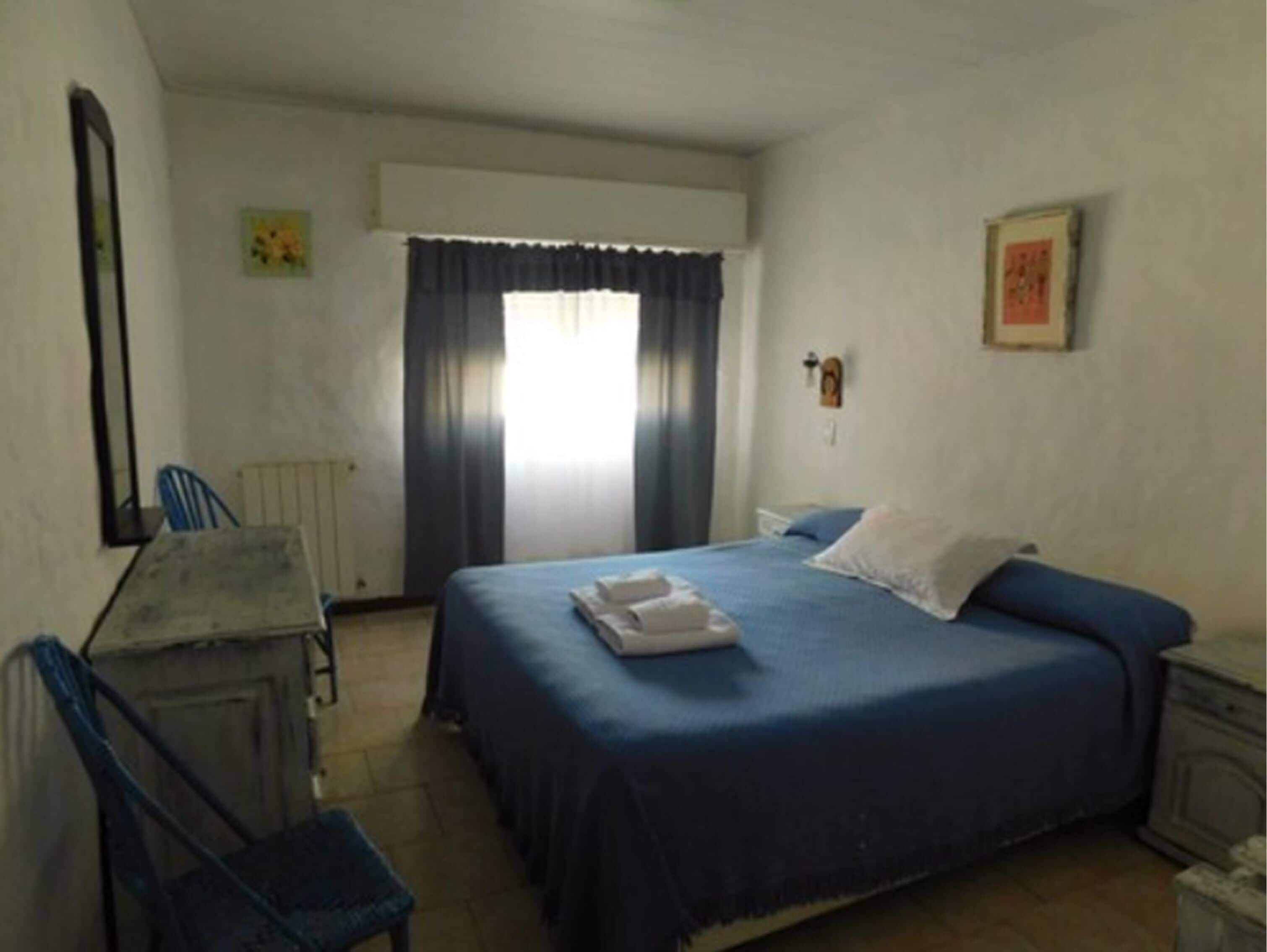 A simple, clean room with a bed, a chair, a desk, and a window with curtains.