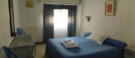 A simple, clean room with a bed, a chair, a desk, and a window with curtains.