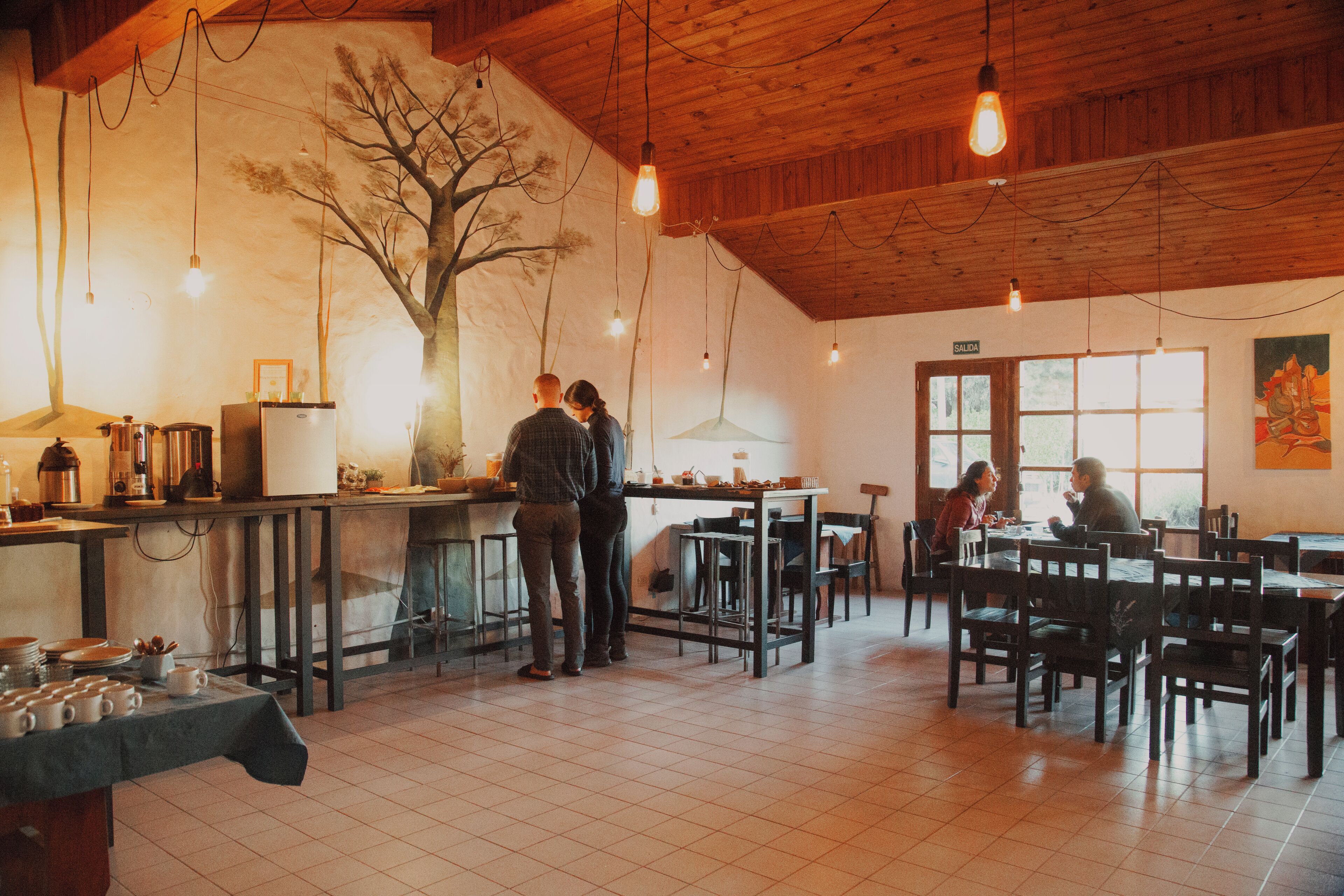 A restaurant with a wooden ceiling, tiled floor, and a mural of a tree. There are tables and chairs for dining, and a counter with a coffee machine.