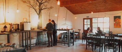 A restaurant with a wooden ceiling, tiled floor, and a mural of a tree. There are tables and chairs for dining, and a counter with a coffee machine.