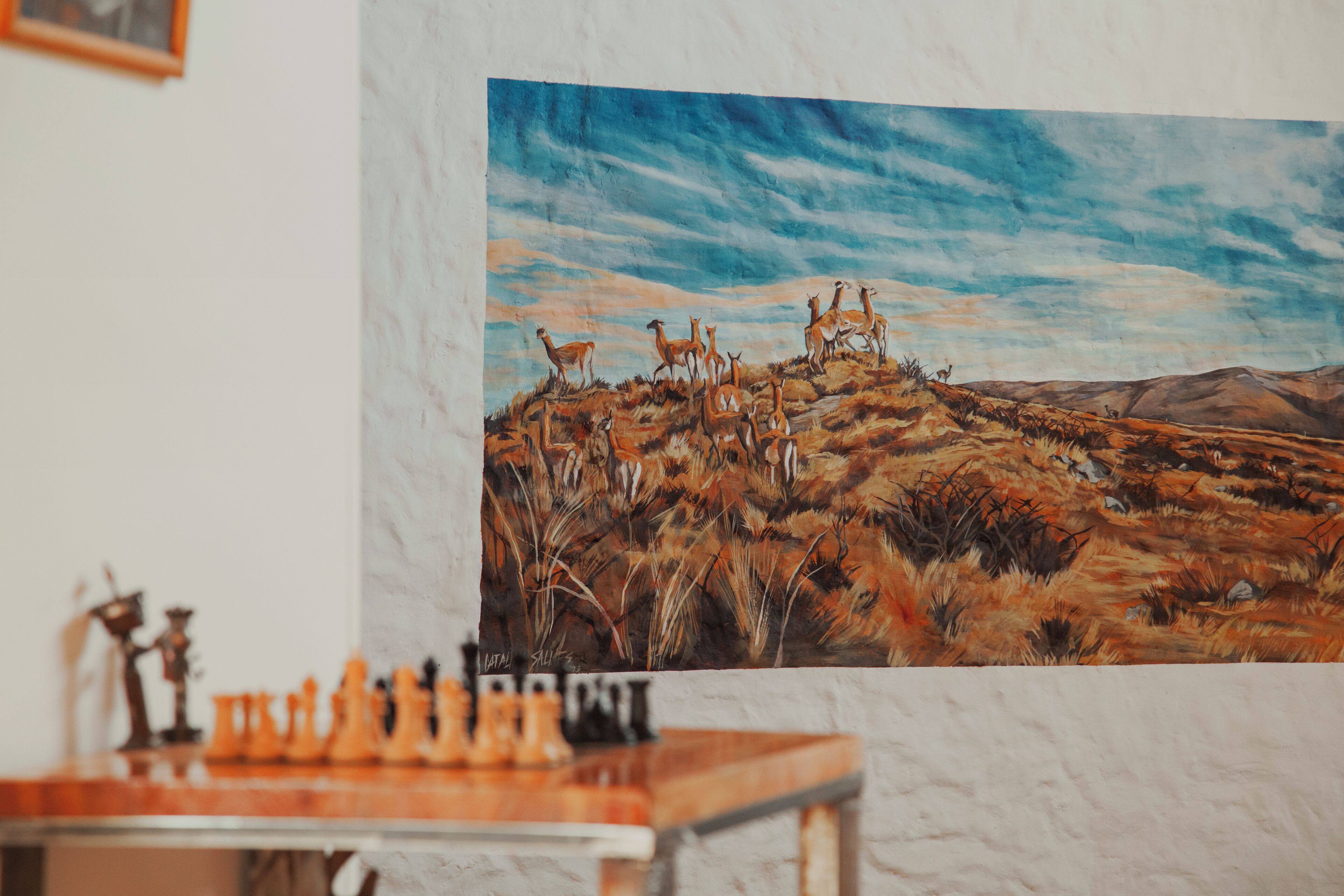 Quarto com uma grande pintura de paisagem desértica na parede, um tabuleiro de xadrez sobre a mesa e uma lareira de mármore.