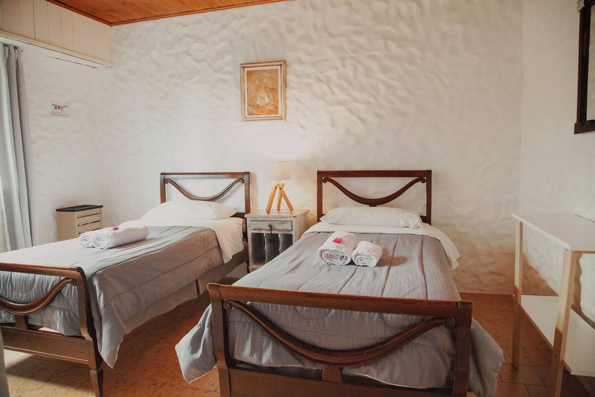 Une chambre avec deux lits en bois, chacun muni d’un oreiller et d’une serviette enroulée. On y trouve une petite table de chevet avec une lampe et un tableau encadré accroché au mur.