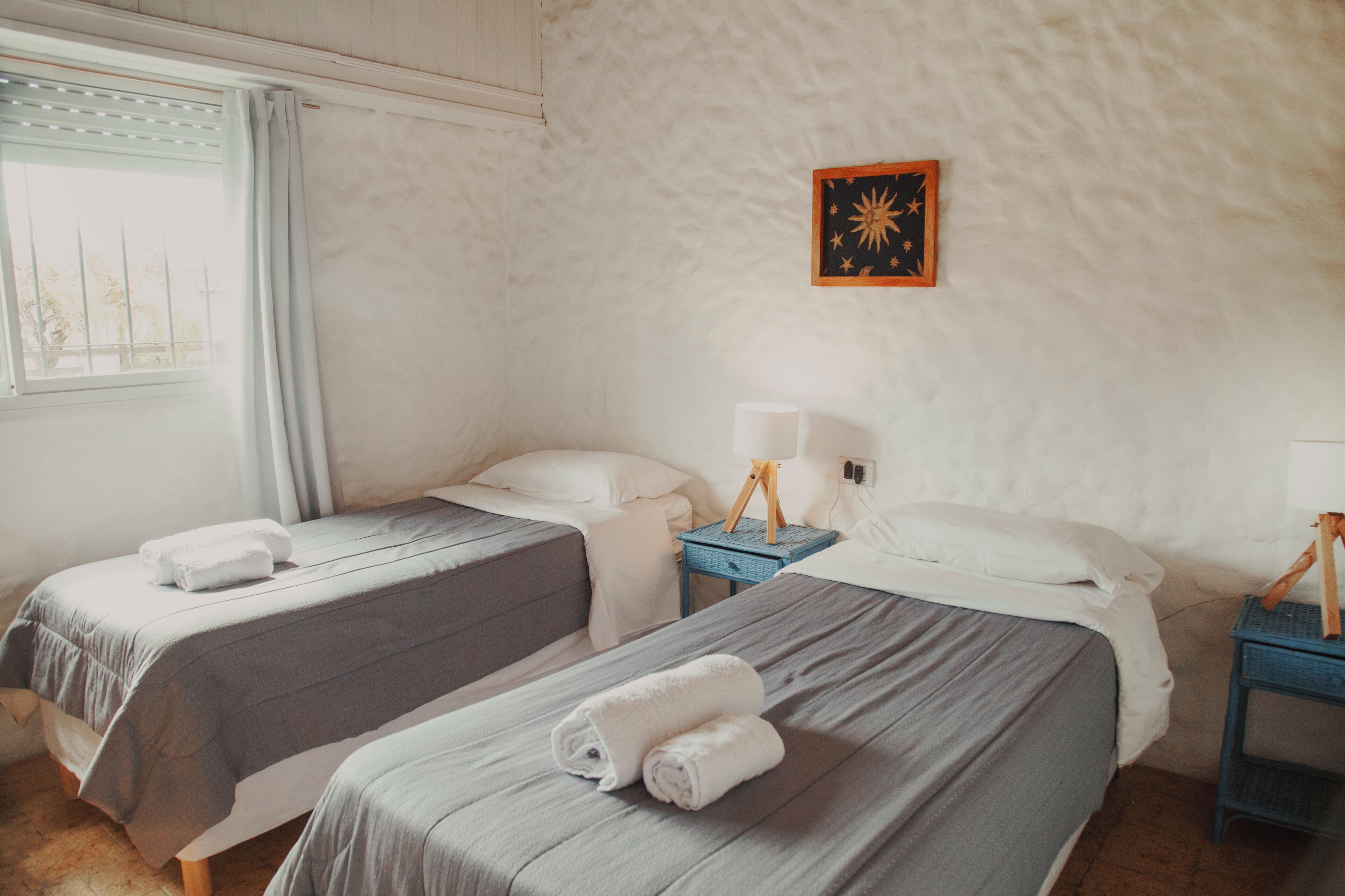 A room with two beds, a window with curtains, a wall-mounted picture, and a nightstand with a lamp.