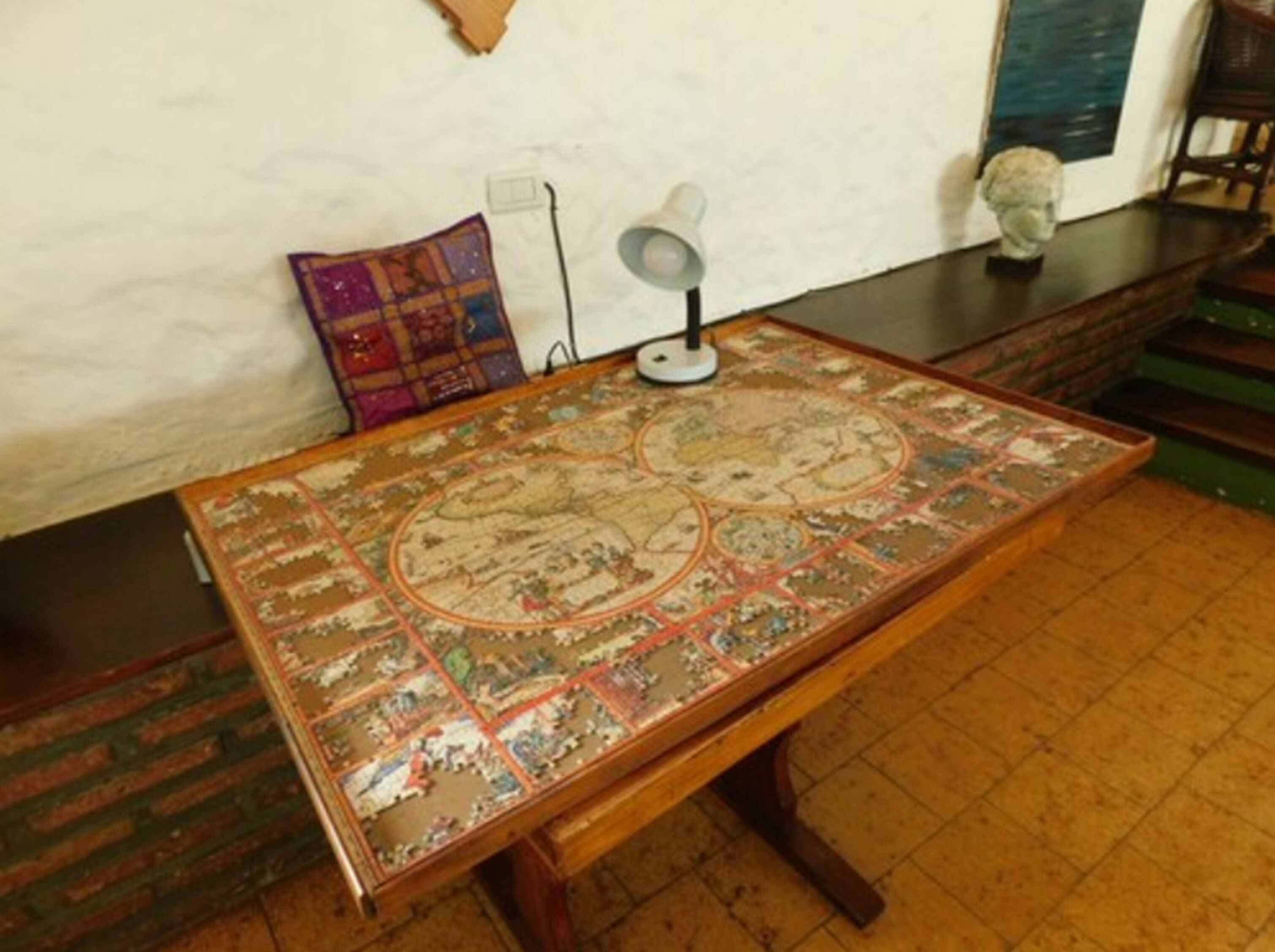A wooden table with a detailed map, a lamp, and a bust on a shelf.