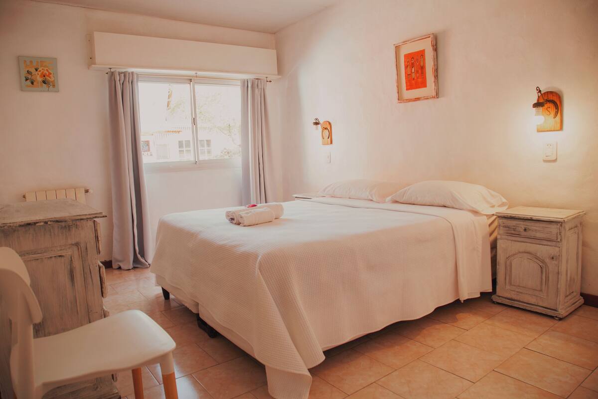 Une chambre à coucher comprenant un lit, une chaise, une table de chevet, une commode et une fenêtre avec des rideaux.