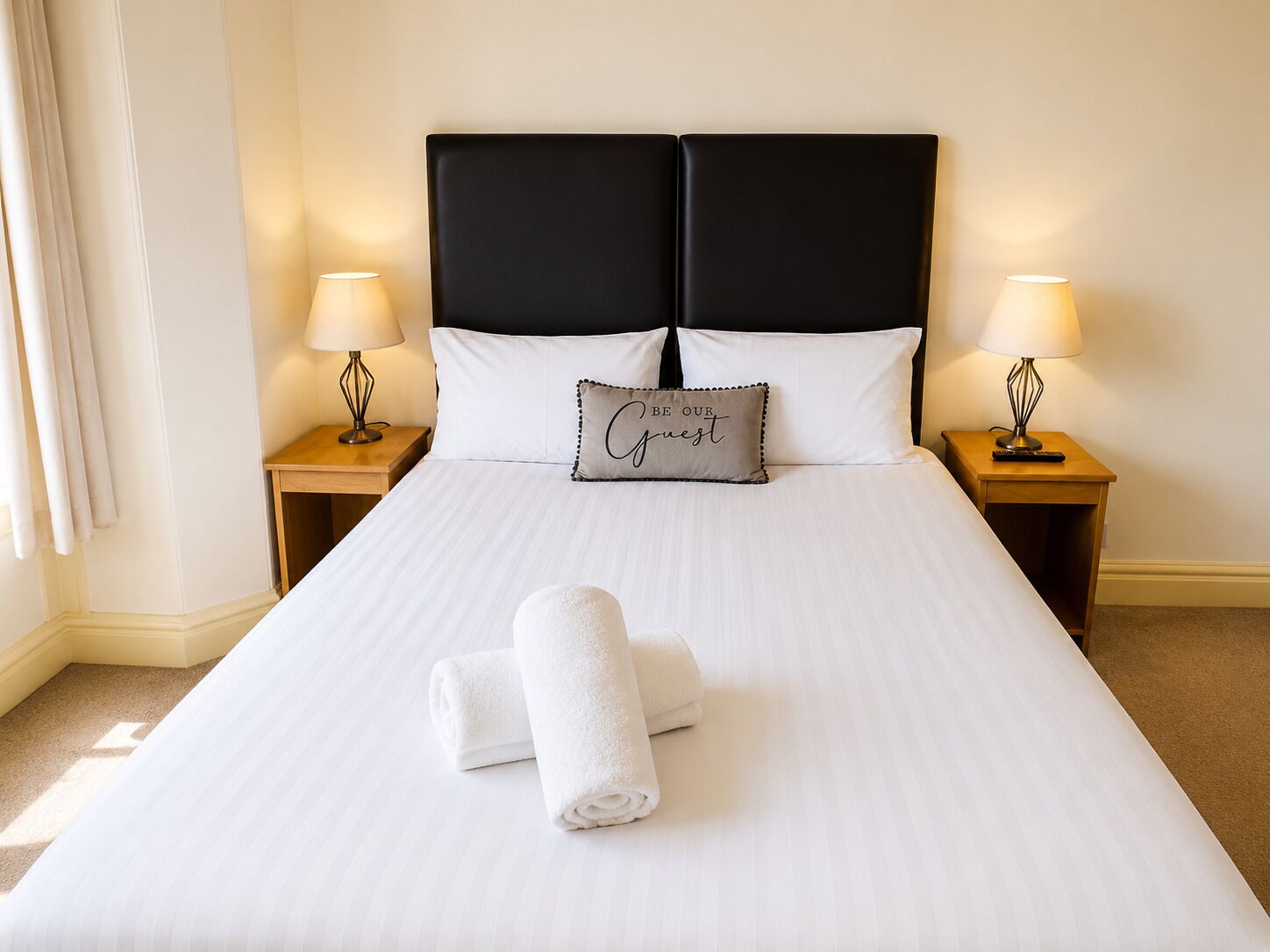 Een net opgemaakt bed met witte lakens, twee bedlampen, een kussen met tekst en een opgerolde handdoek op het bed.