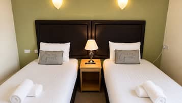 Ein Hotelzimmer mit zwei Betten, jedes mit einem Kopfkissen mit der Aufschrift 'BE OUR Guest', einem Nachttisch mit einer Lampe und einer Wandleuchte.