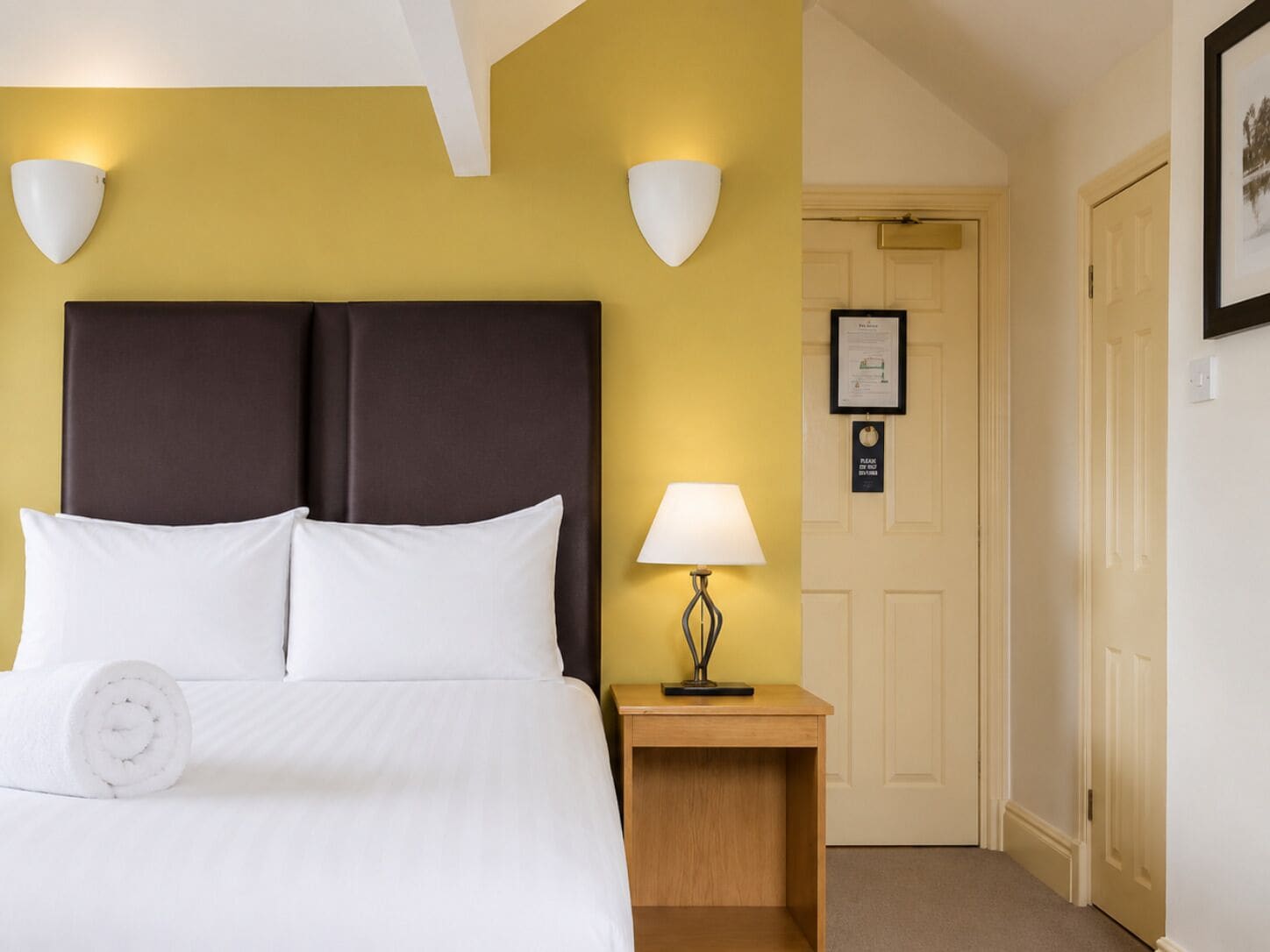 A hotel room with a bed, a nightstand with a lamp, and a door with a framed notice.