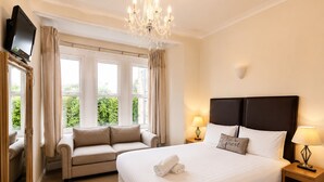 A hotel room with a large bed, a sofa, a TV, a chandelier, and a view of greenery. - Heath Cottage Hotel (Dewsbury)