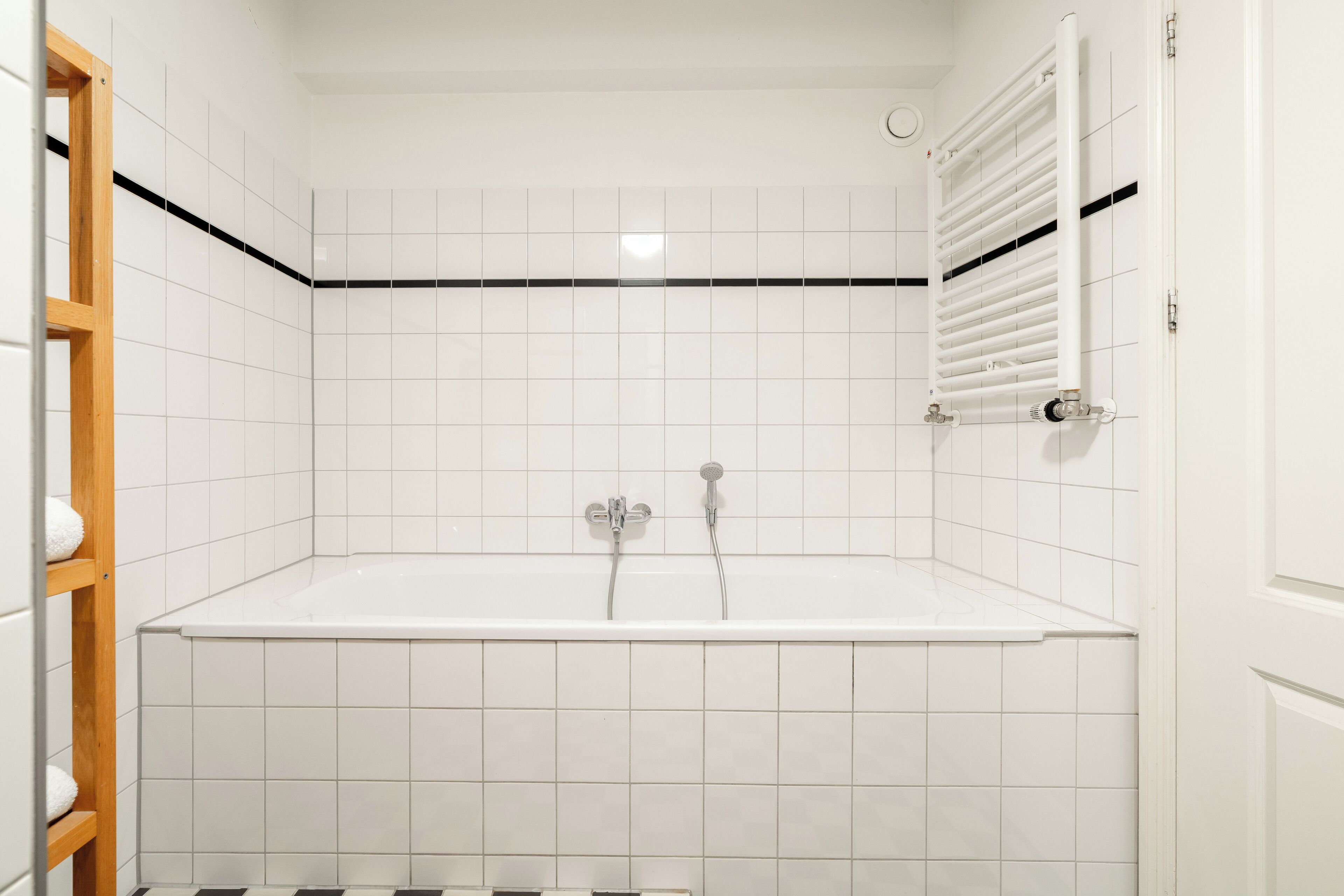 Un baño con paredes de azulejos blancos, una bañera, ducha y un estante de madera con toallas.