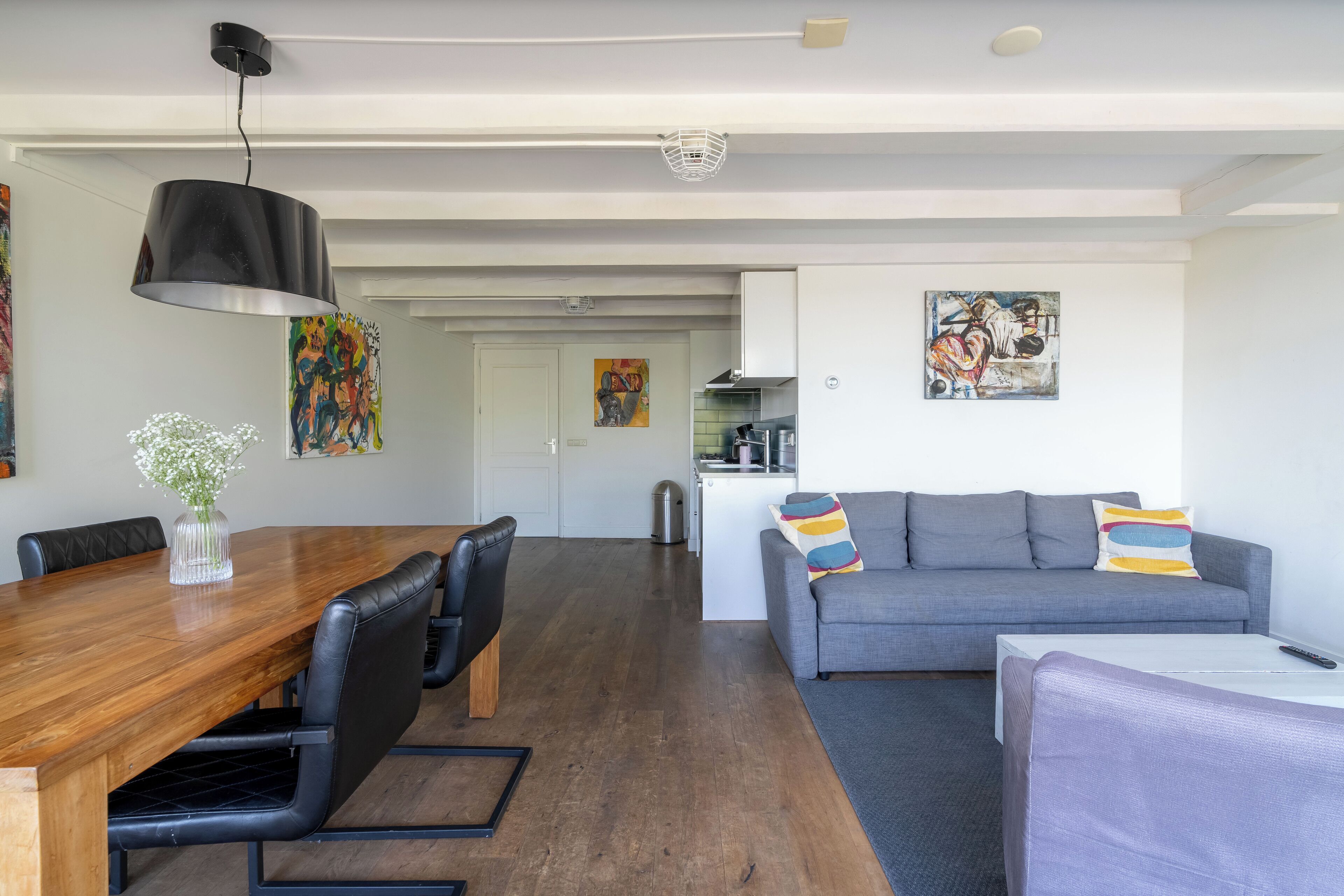 A modern living room with a wooden dining table, black leather chairs, a grey sofa, and a white coffee table. There is a kitchen area in the background with a white cabinet and a painting on the wall.