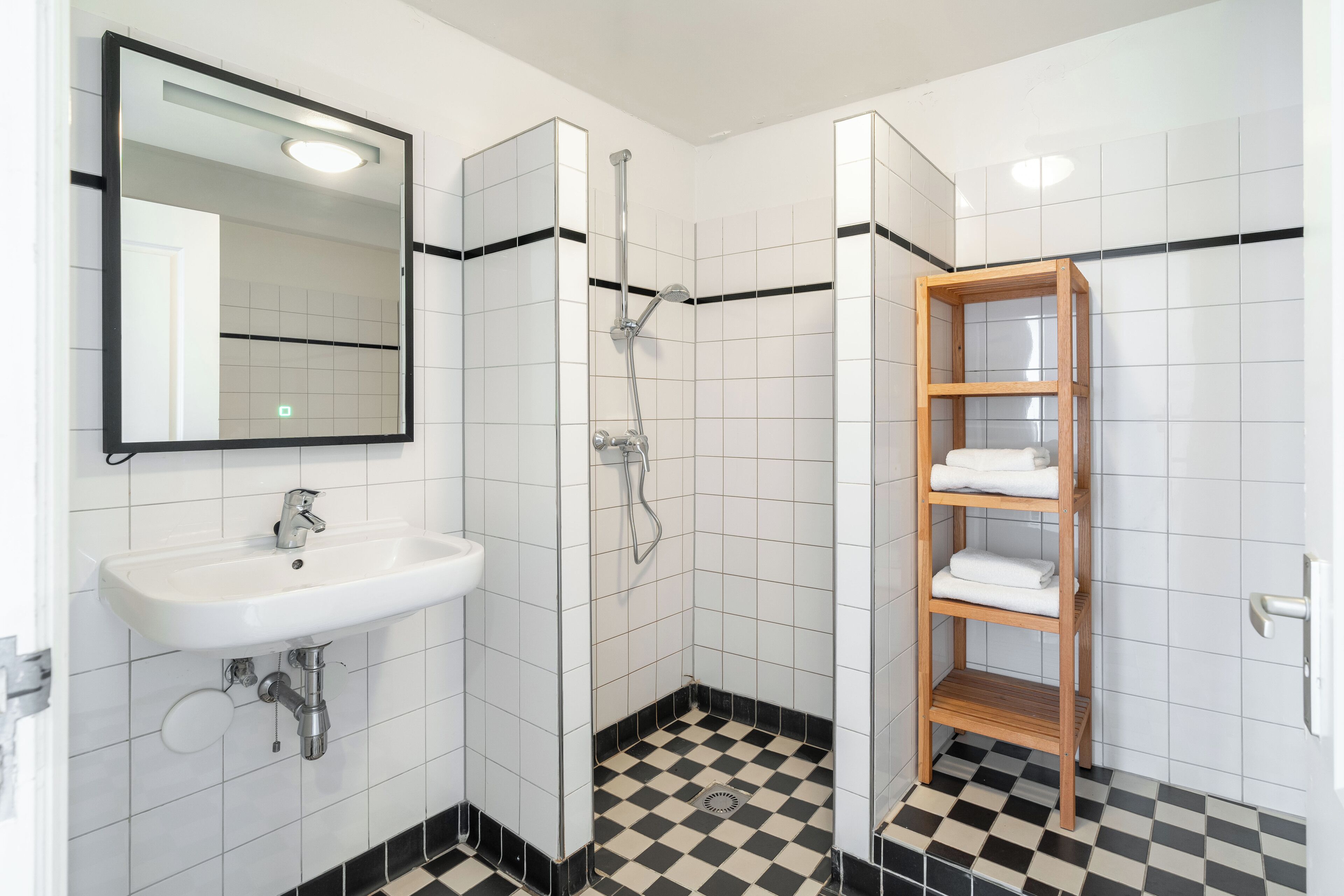 A bathroom with a white sink, a mirror, a shower, and a wooden shelf with towels.