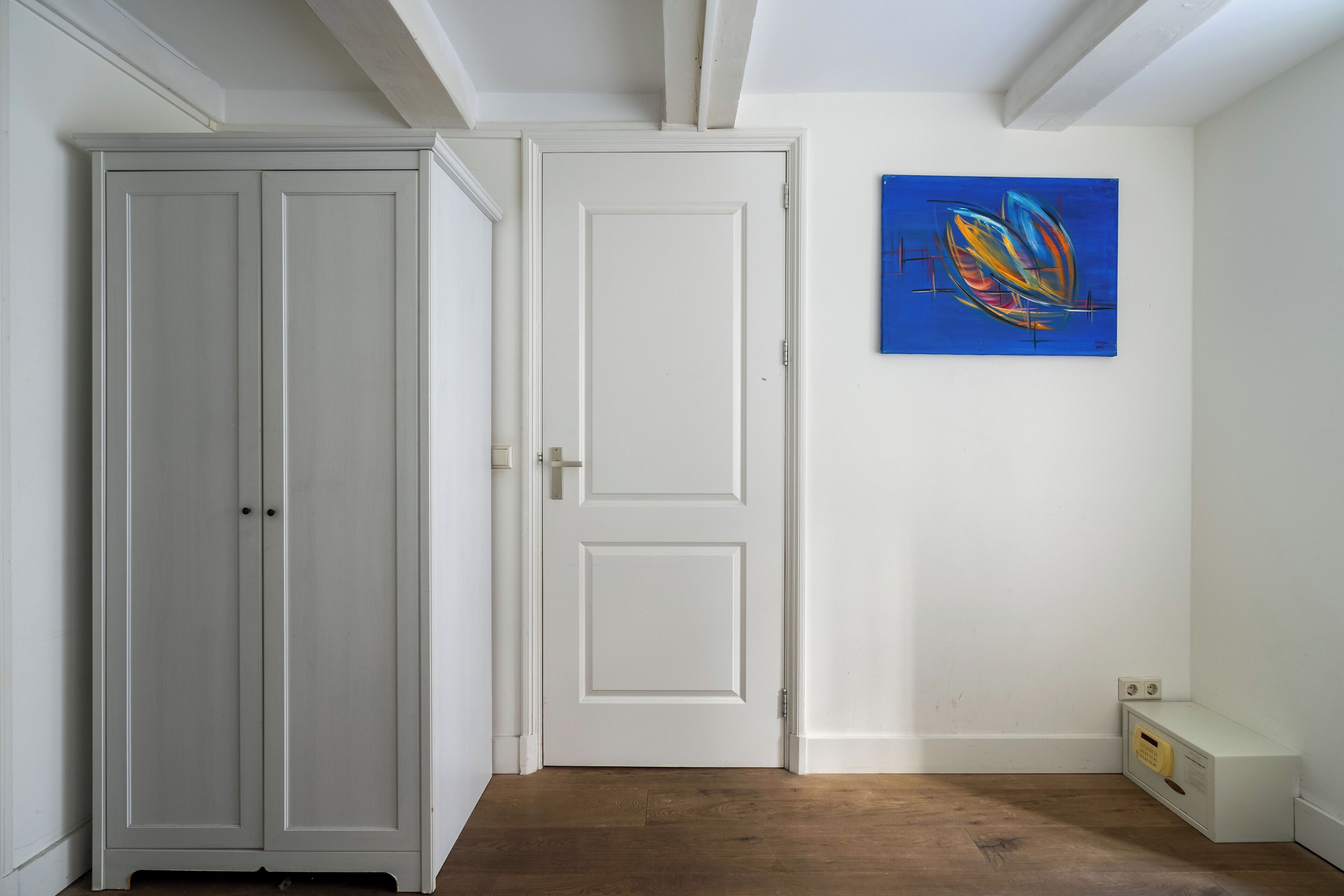 Una habitació amb una porta blanca, un quadre abstracte blau a la paret i un terra de fusta.