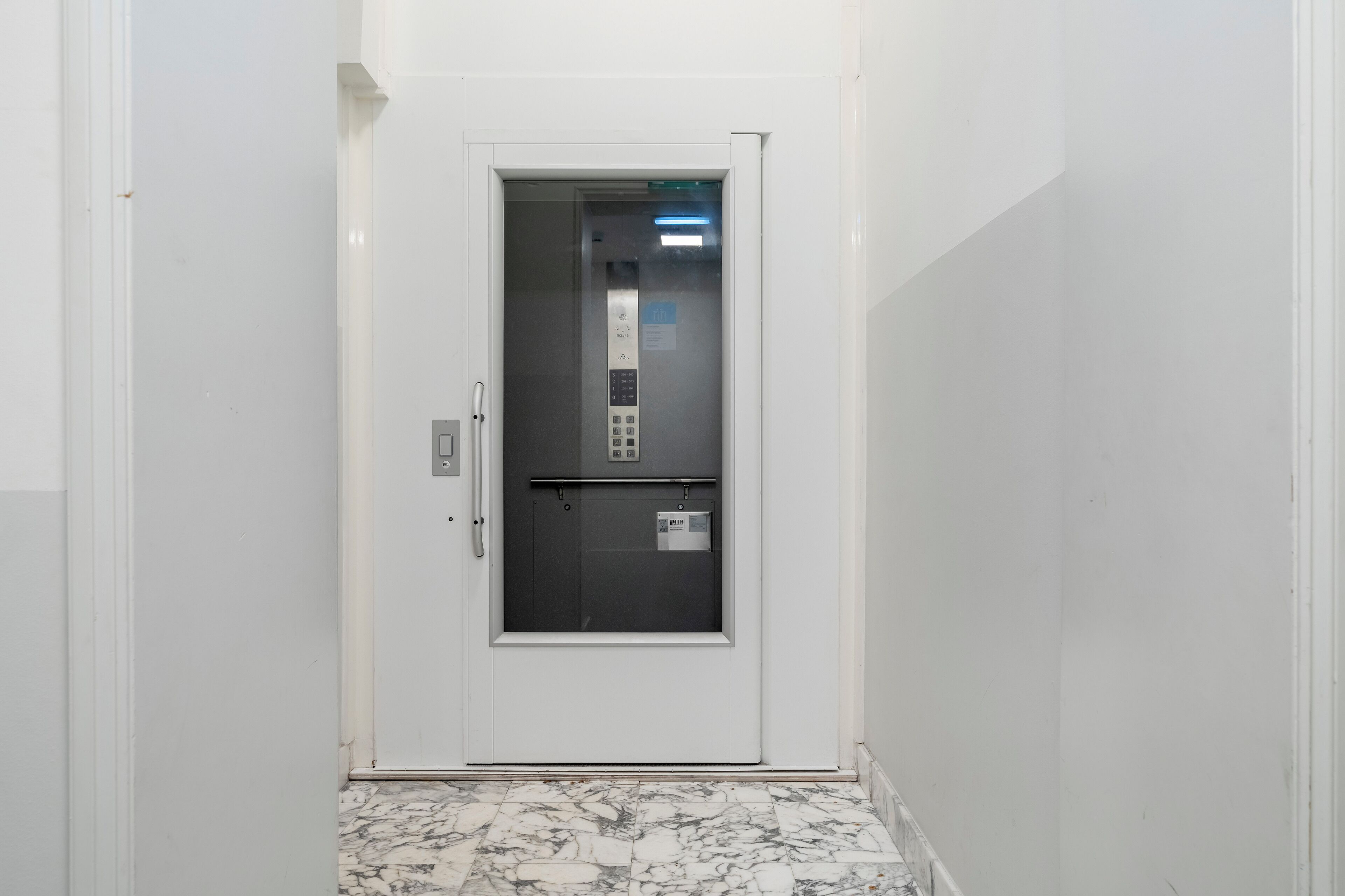 Un couloir étroit avec une porte d’ascenseur blanche et un sol en marbre.