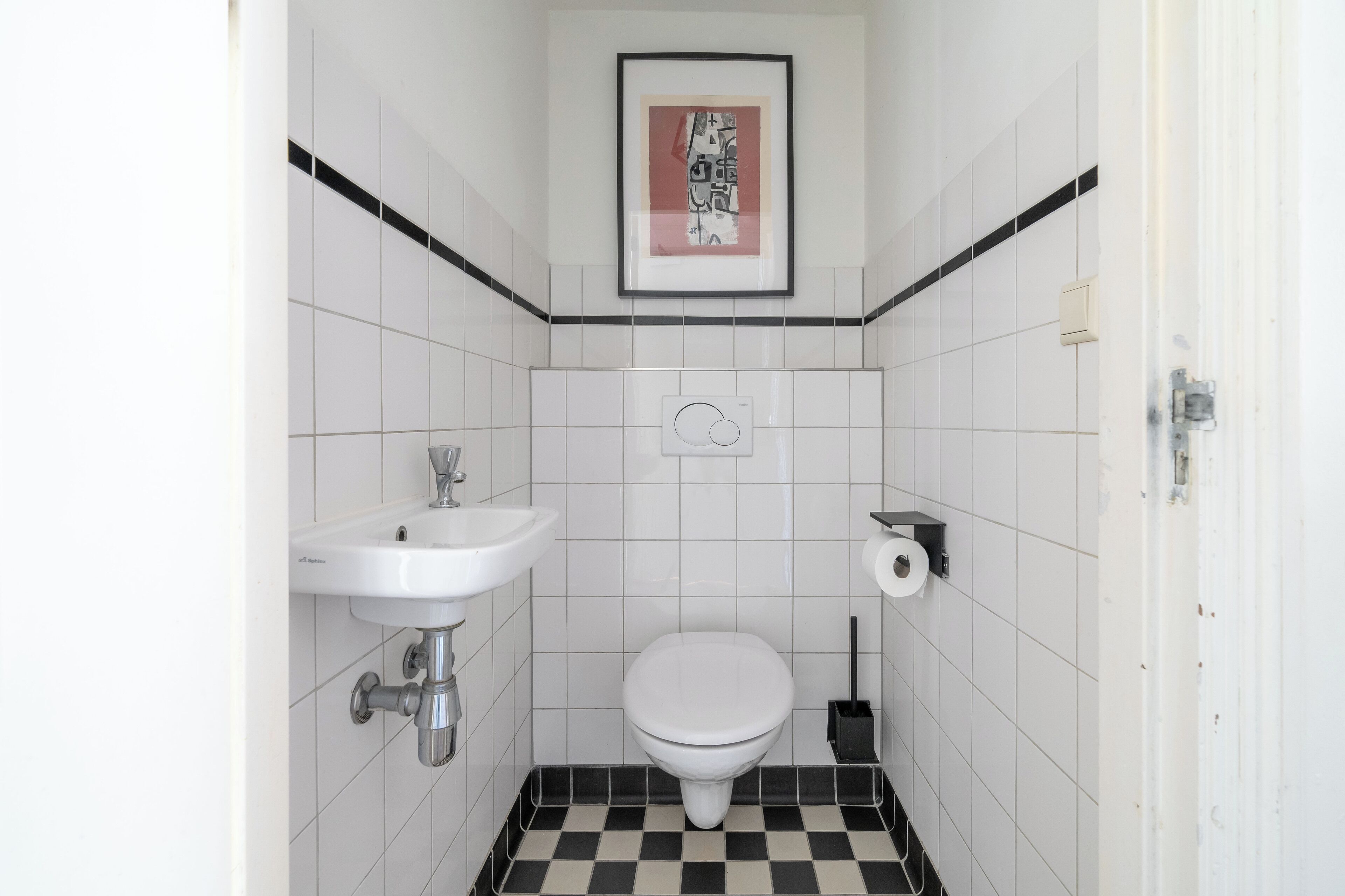 A small, white bathroom with a checkered floor, a white sink, a toilet, and a framed picture on the wall.