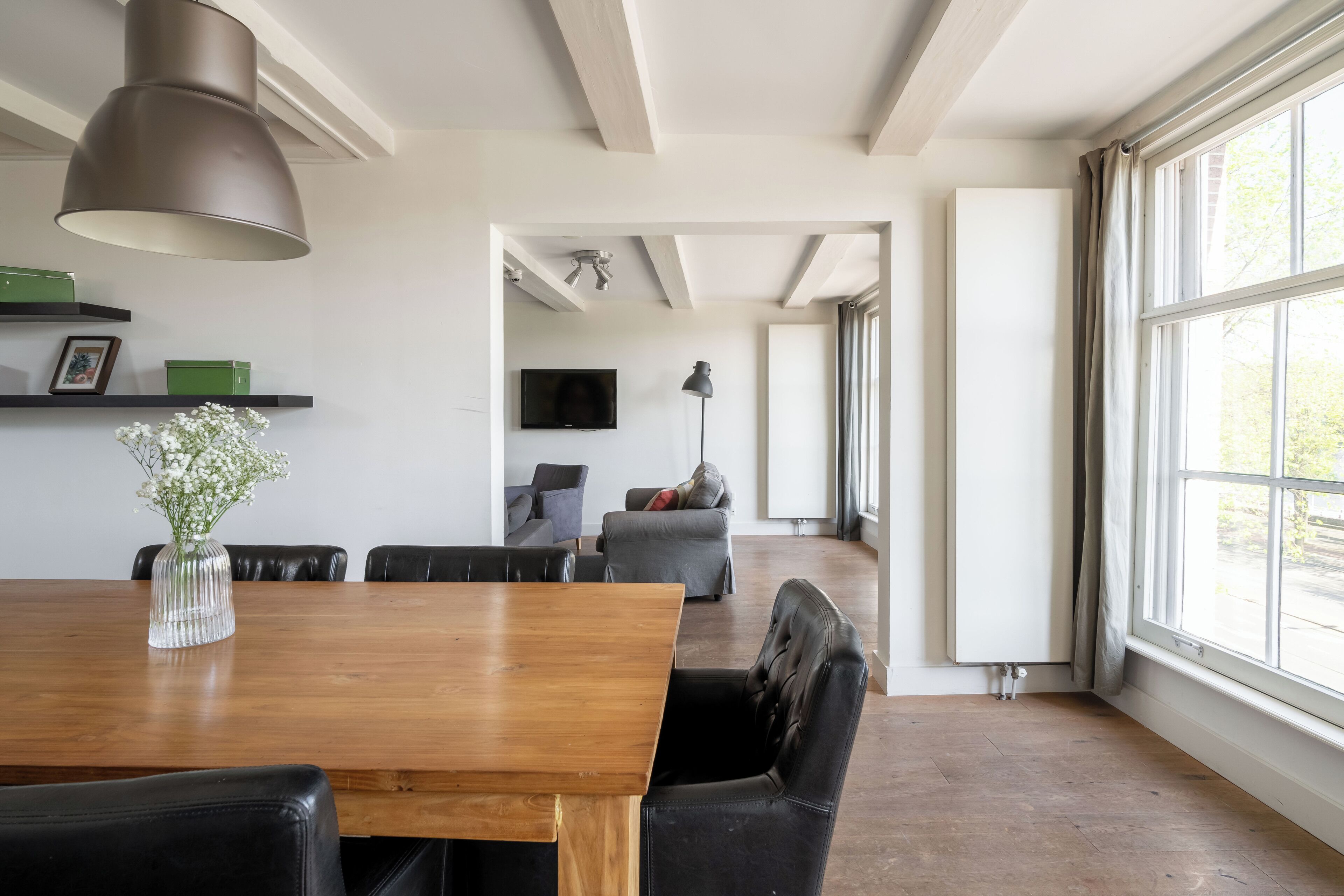 A modern dining area with a wooden table and chairs, a vase of flowers, a large pendant light, a flat-screen TV mounted on the wall, and a view through a large window to the outdoors.