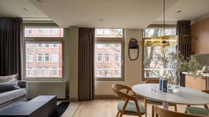 Een moderne woonkamer met een bank, een eettafel met stoelen, grote ramen met gordijnen en uitzicht op gebouwen.
