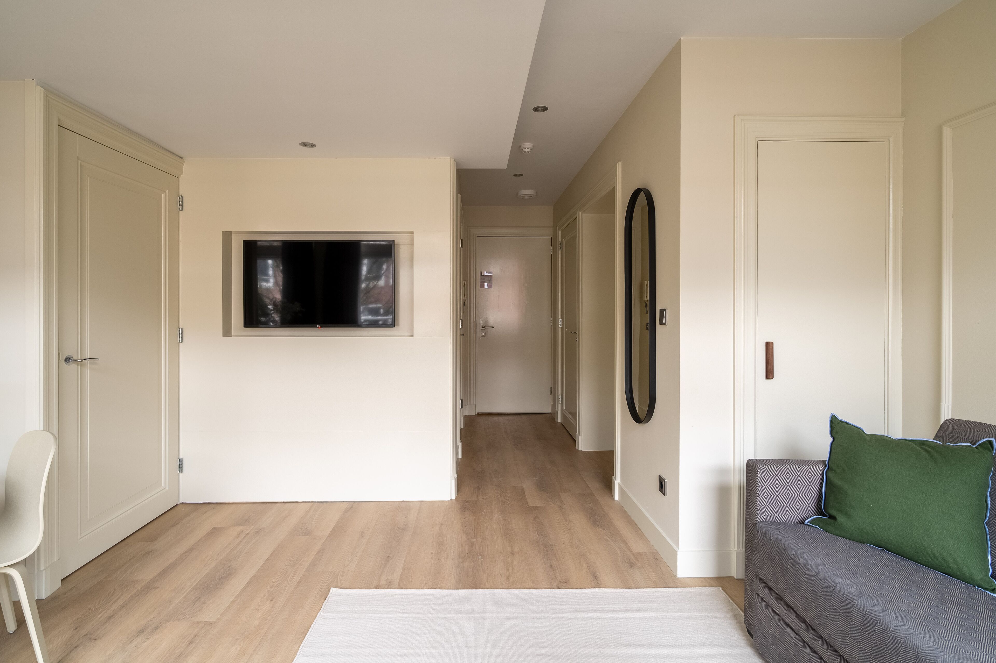 Una sala moderna con piso de madera, un sofá gris con un cojín verde, un sillón blanco y un televisor de pantalla plana montado en la pared.