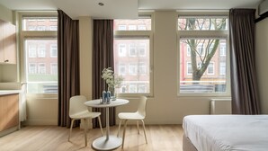 Een hotelkamer met een bed, een bureau, een stoel, een tafel met flessen en grote ramen met gordijnen.
