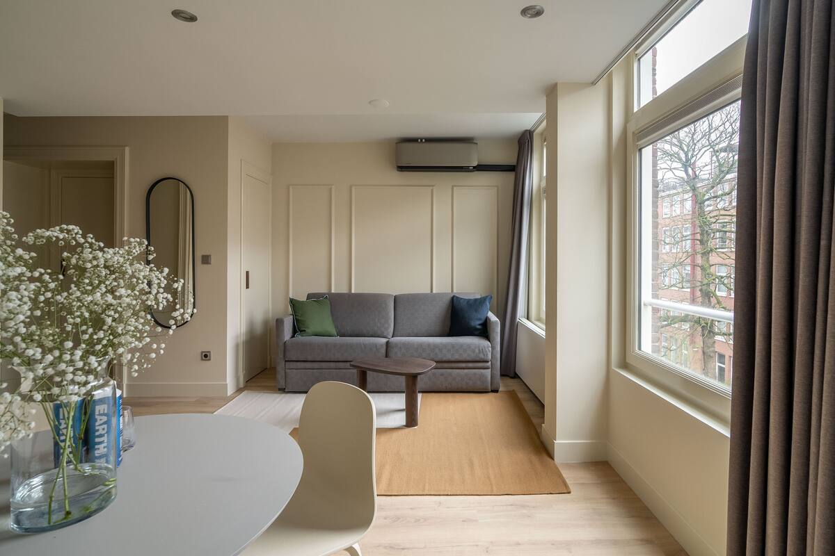 A living room with a sofa, a coffee table, a dining table with chairs, a vase with flowers, a mirror, and a window with curtains.