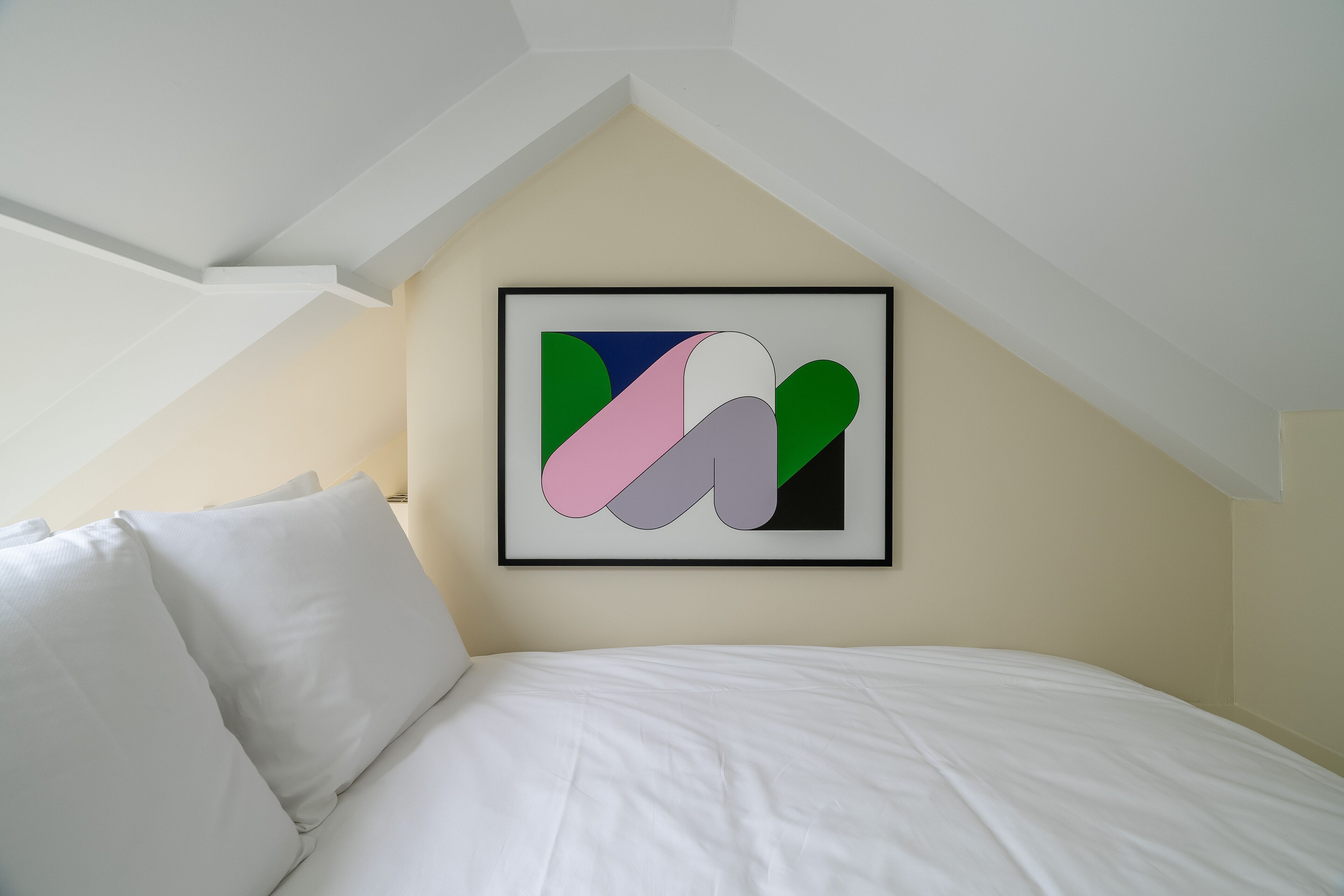 A bedroom with a white bed, a framed abstract painting on the wall, and a sloped ceiling.