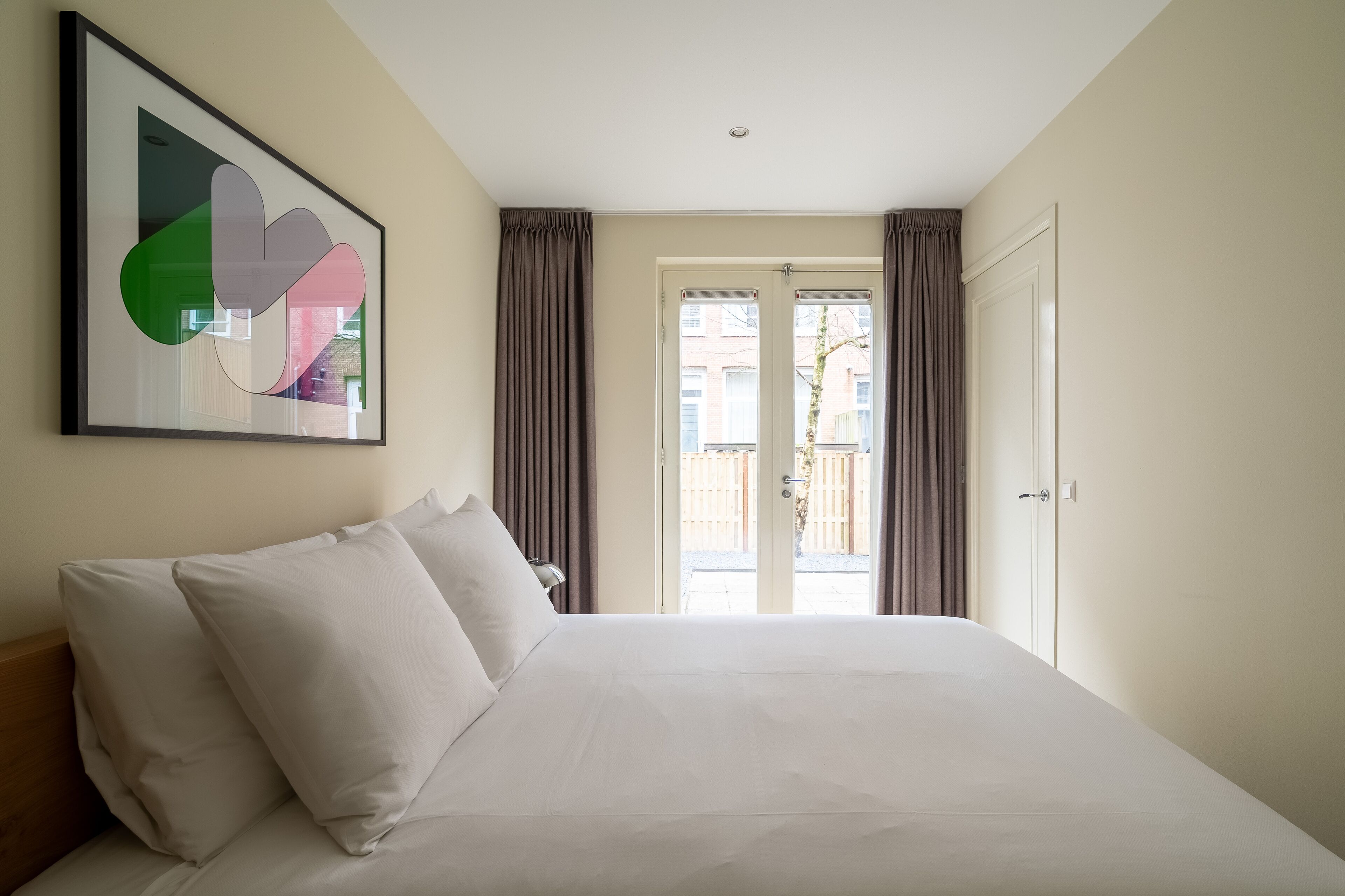A hotel room with a large bed, a framed abstract artwork, and a view through a glass door.