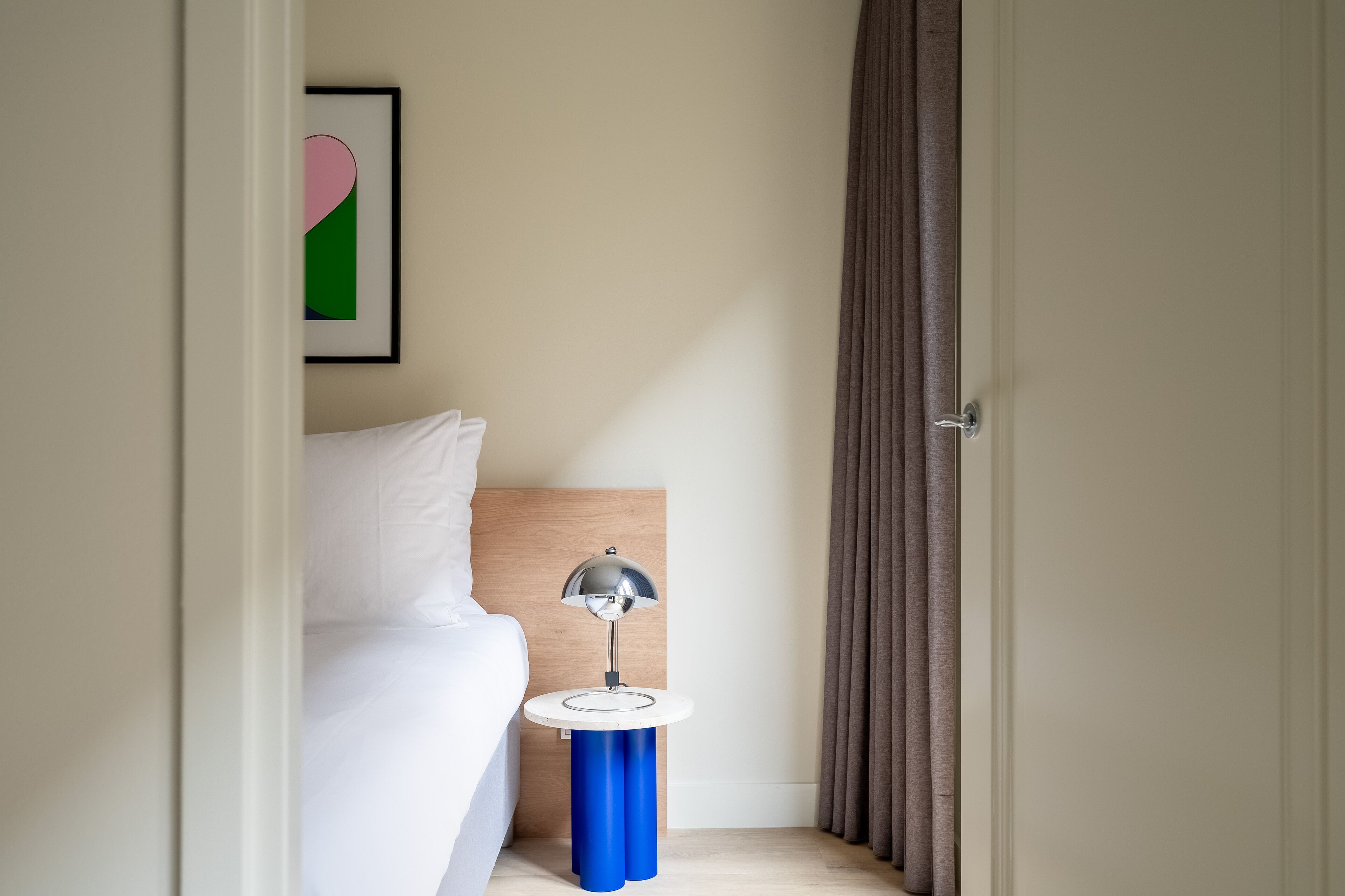 Habitació d'hotel amb un llit, una taula de nit amb una làmpada, un quadre amb marc a la paret i una porta amb cortines.