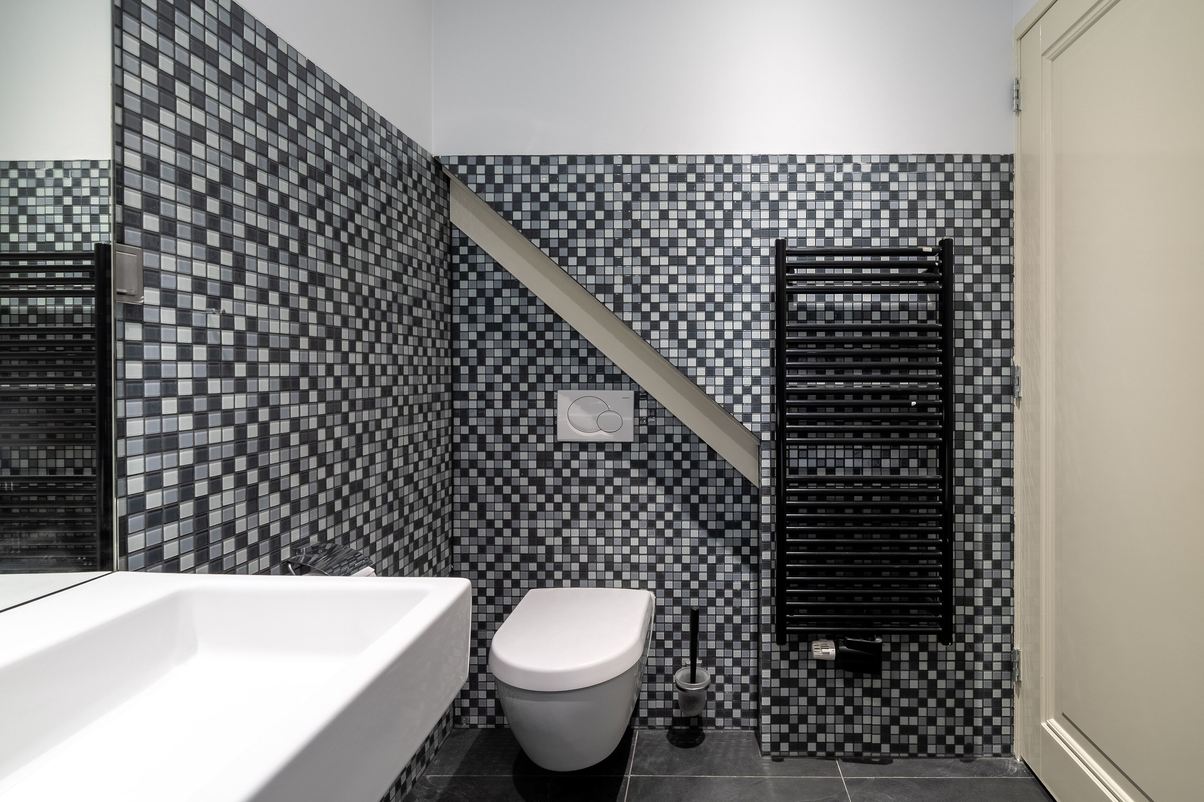 Un baño con pared de mosaicos, un inodoro, un toallero negro y una bañera blanca.