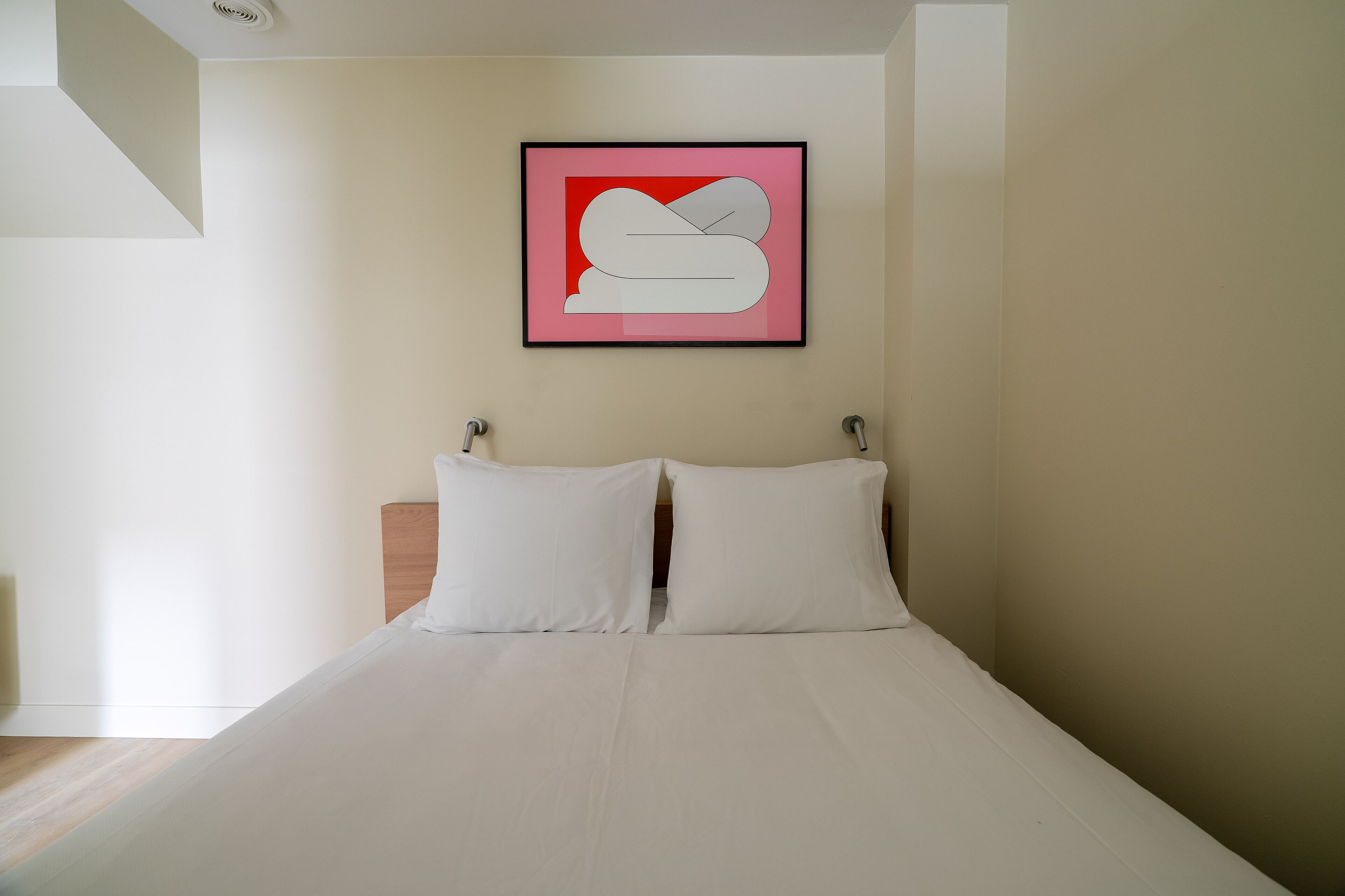 Una cama bien hecha con ropa de cama y almohadas blancas, una obra de arte abstracta enmarcada en la pared y un cabecero de madera.