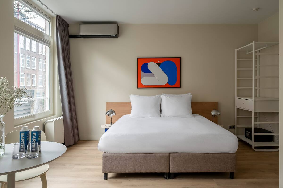 A modern hotel room with a large bed, a side table with two bottles, a lamp, and a view of buildings through a window.