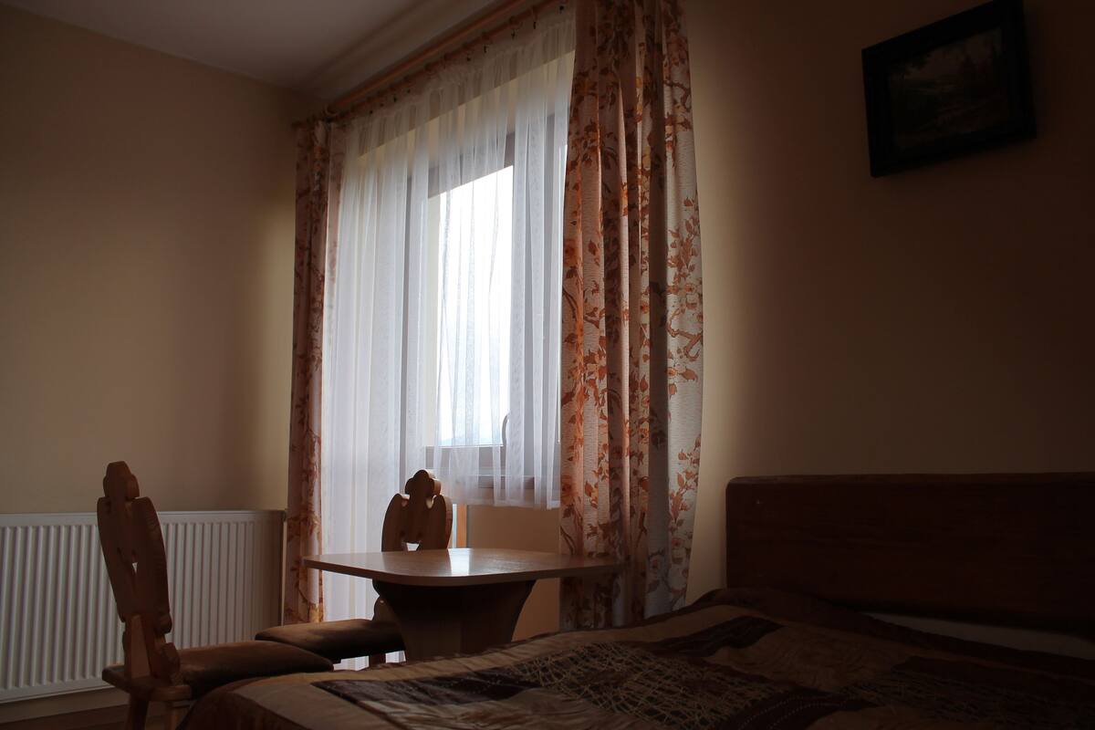 Une chambre à coucher avec un lit, une chaise en bois, une petite table et une fenêtre ornée de rideaux légers.