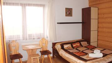 Un dormitorio con cama, un armario de madera, una mesita con sillas y una ventana con cortinas.