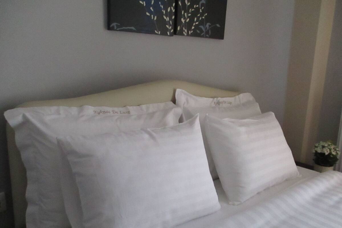 Een netjes opgemaakt bed met witte linnens en kussens, een crème-kleurig hoofdeinde, en een kleine potplant op het nachtkastje.